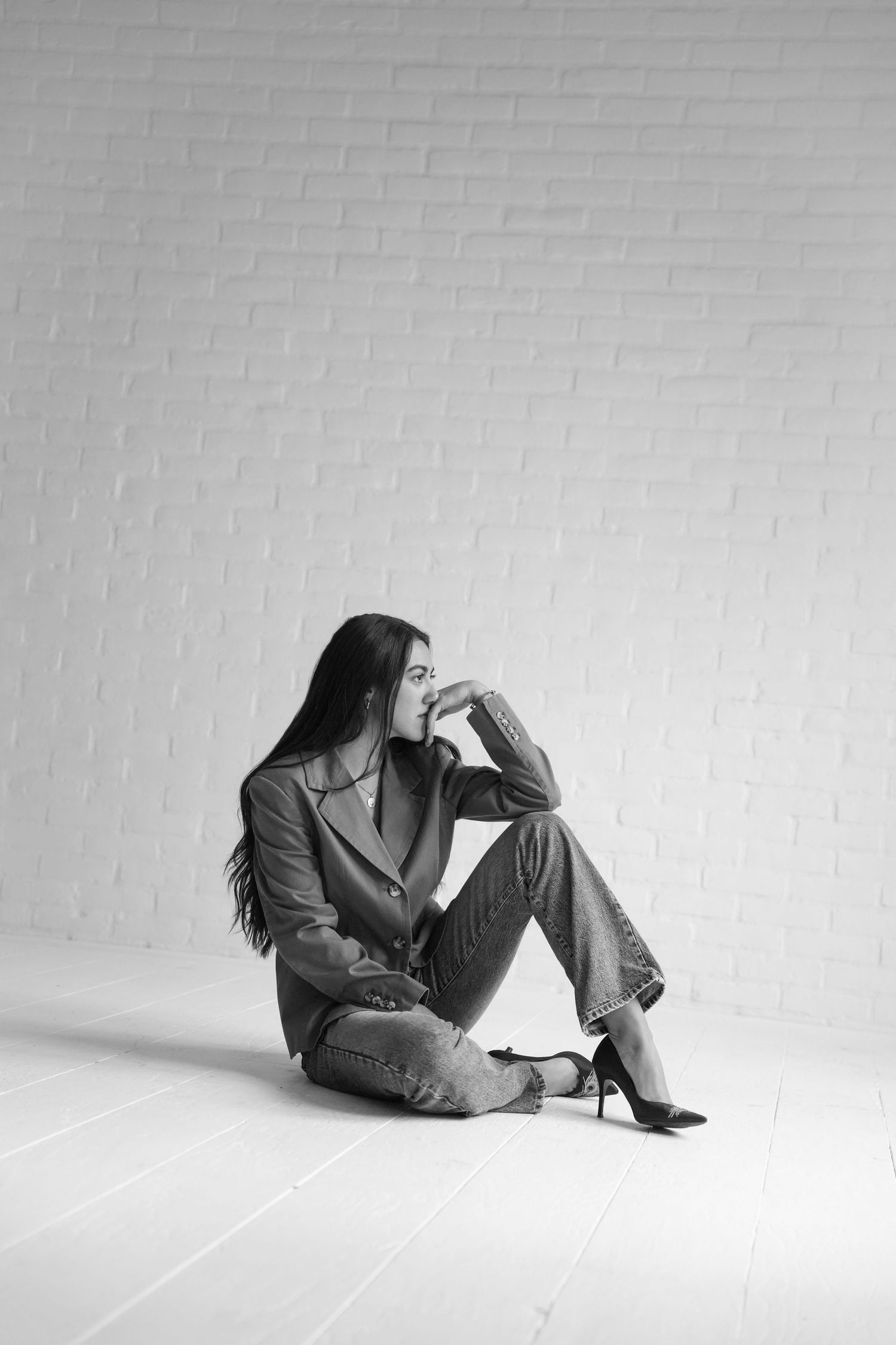 Black and white photo of a stylish woman in blazer and jeans sitting on floor against white brick wall.