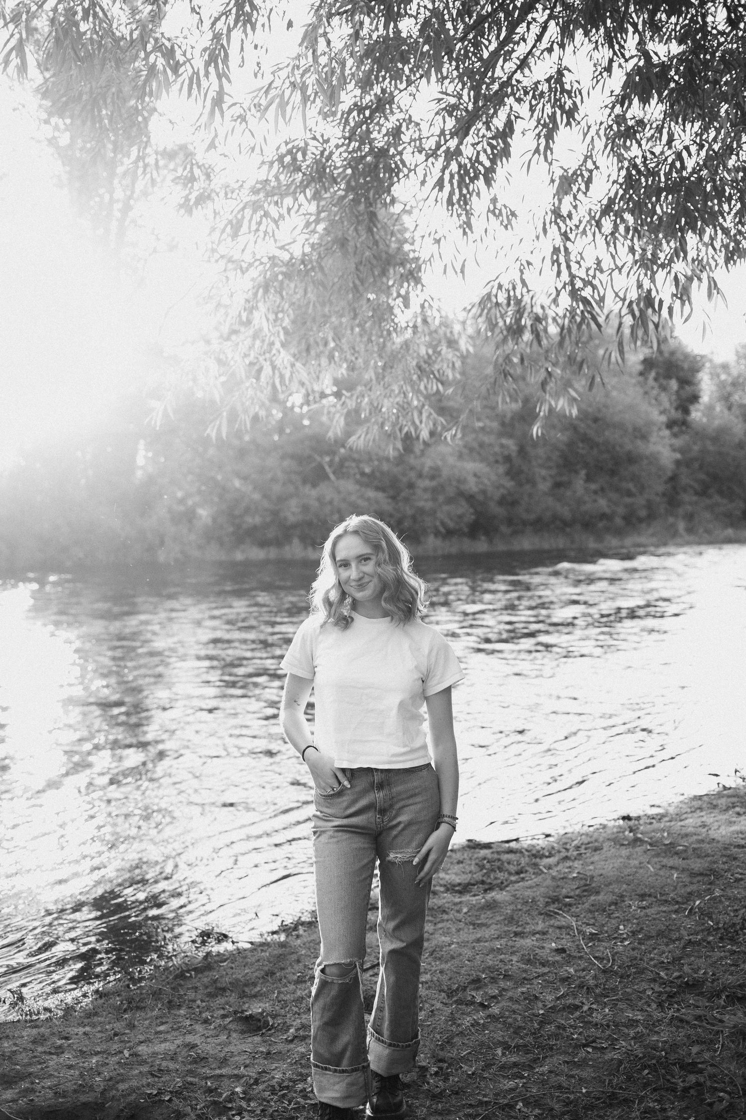 Black and white portrait of a woman in jeans and tee smiling by a misty sunlit riverside.