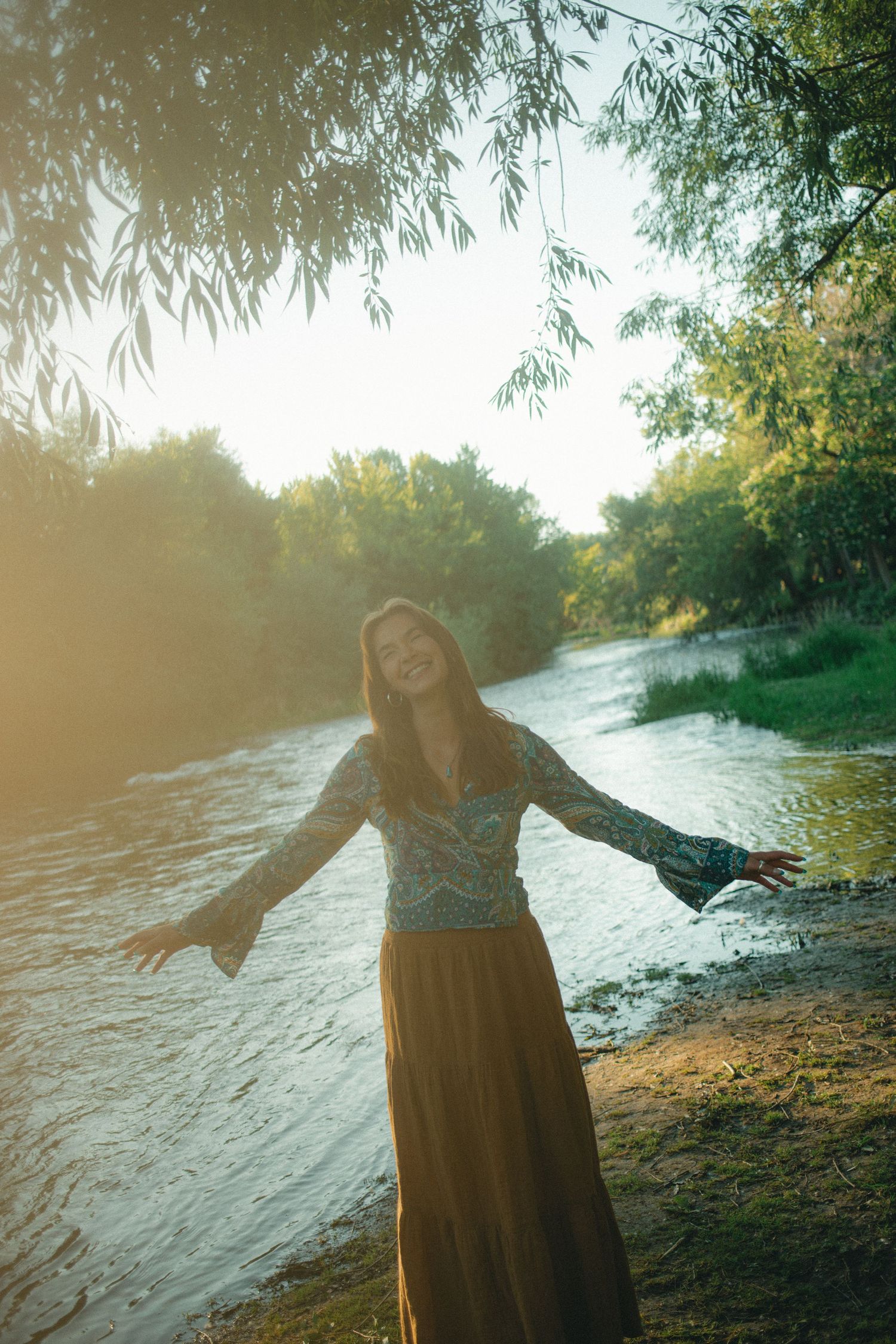 Joyful woman in floral blouse and brown skirt with arms outstretched by a glowing sunlit riverside.