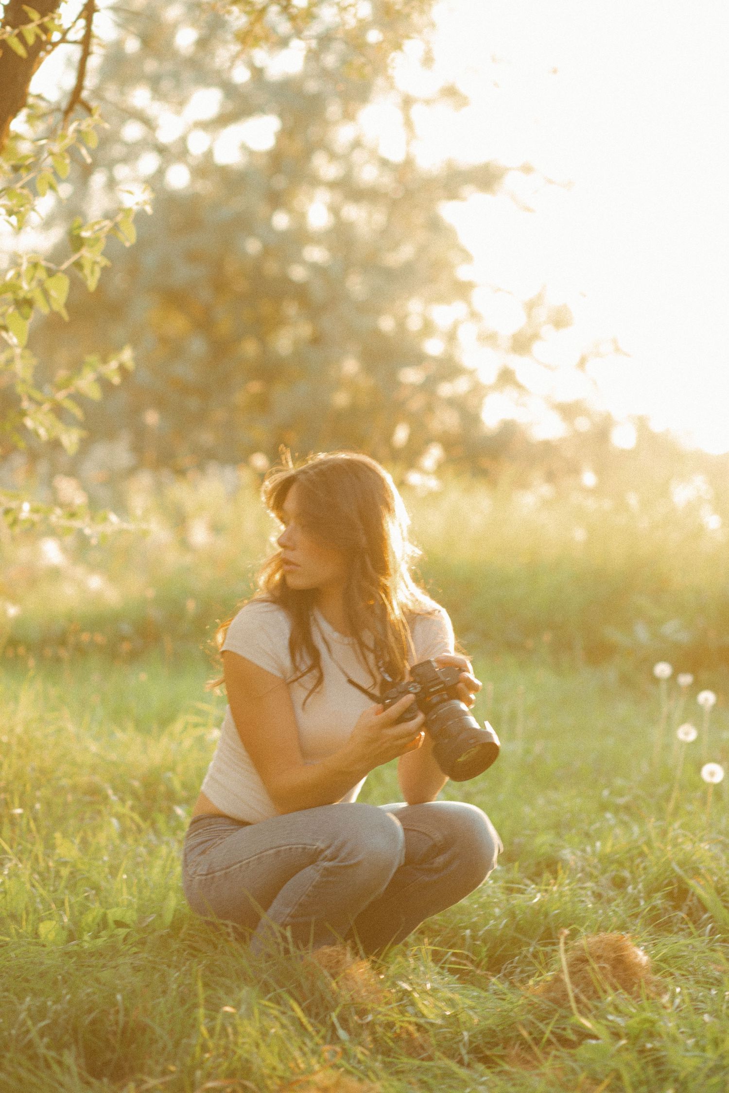 Woman crouching in sunlit grass holding a DSLR camera, surrounded by trees with golden backlight.