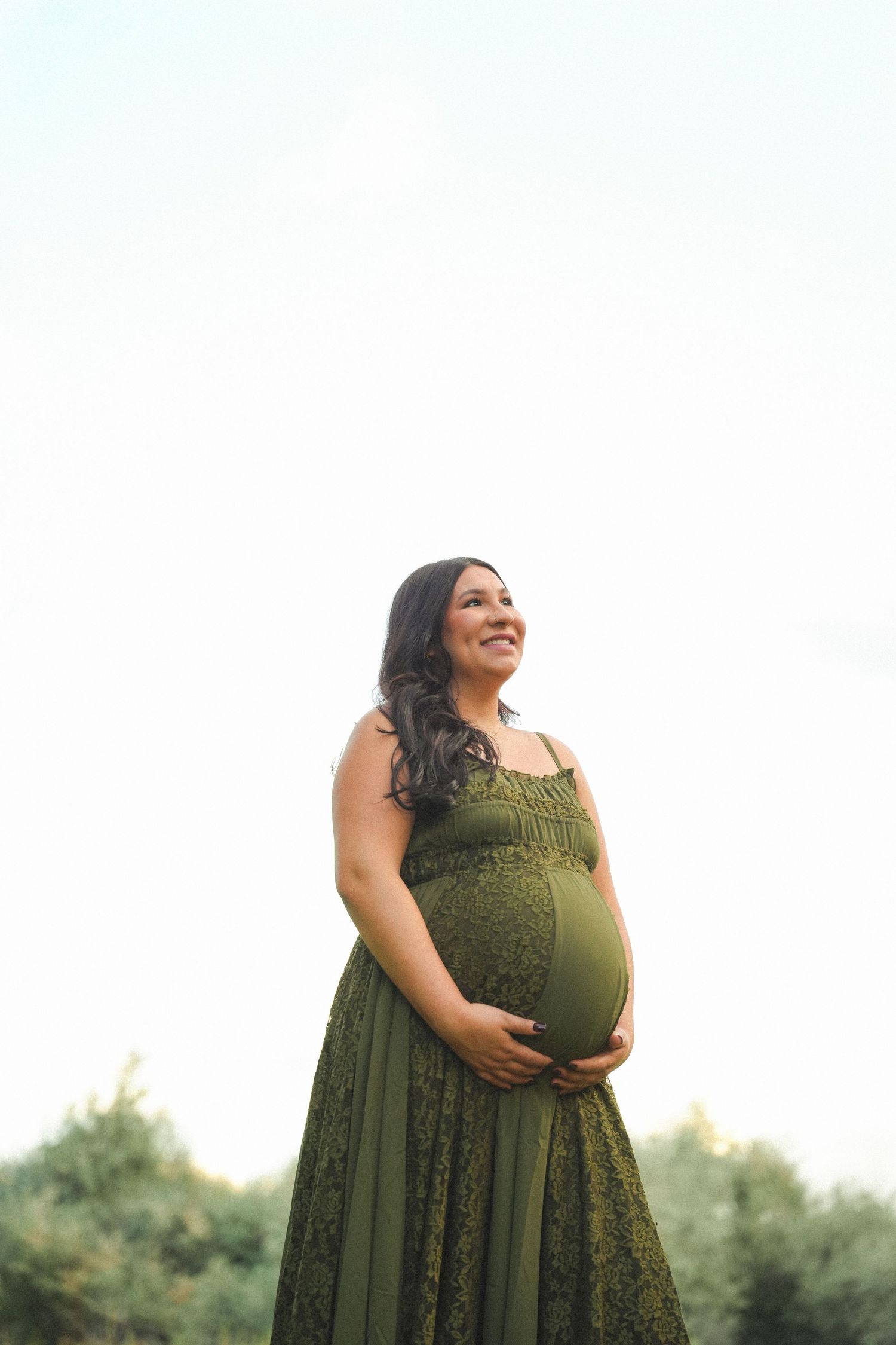 Smiling pregnant woman in olive green lace dress cradling her baby bump outdoors against a bright sky.