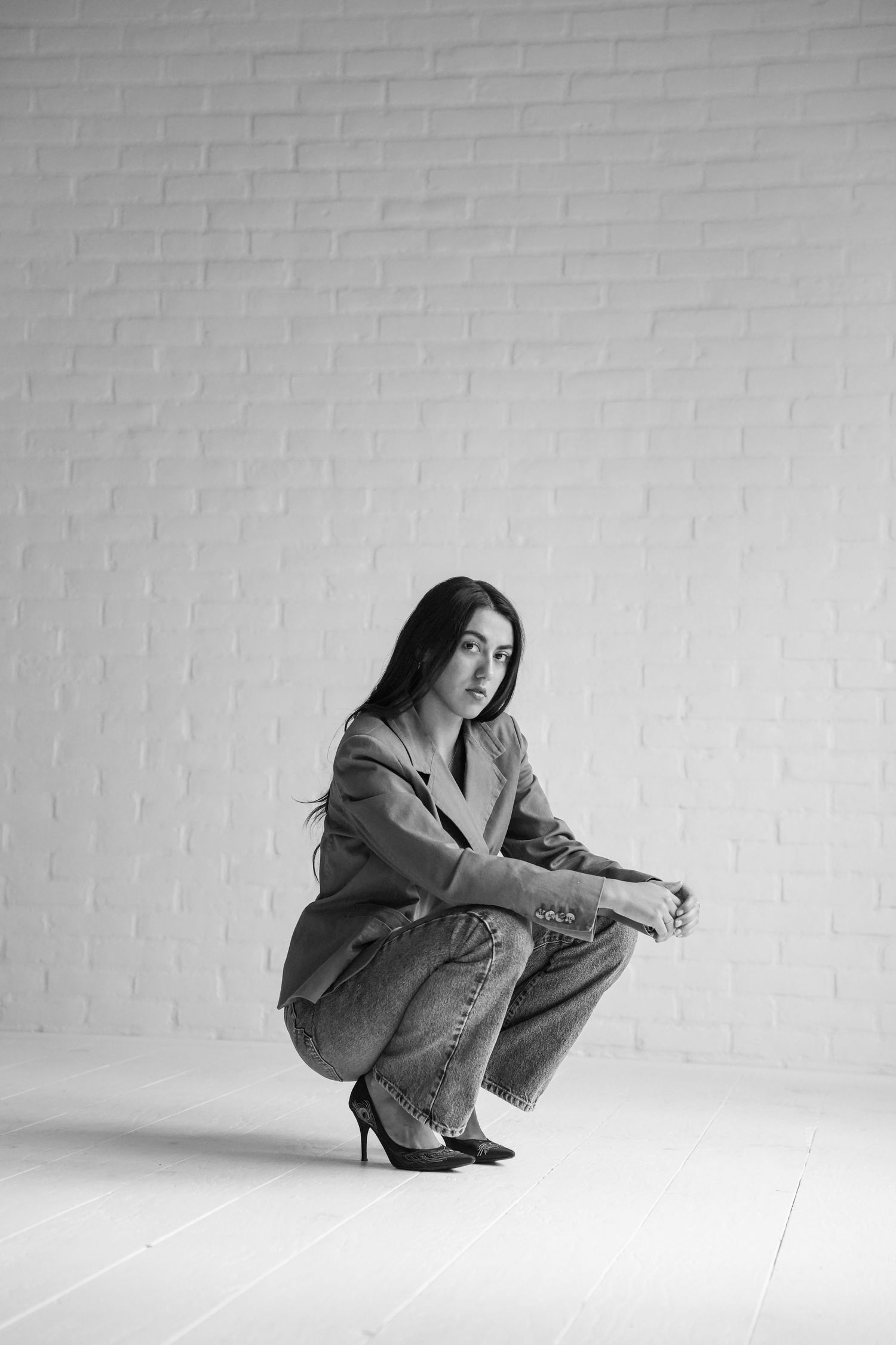 Black and white photo of a young woman crouching in wide-leg jeans, blazer, and heels against a white brick wall.