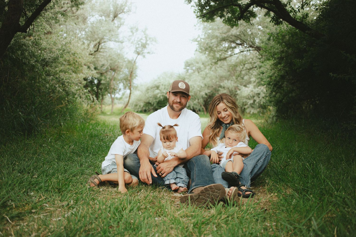 Happy family of five sitting together on grass path surrounded by lush green trees during outdoor photo session.