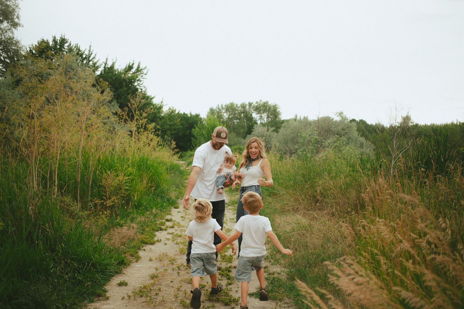 Family of five walking together on sunny dirt path surrounded by tall grass, parents holding hands with young children.