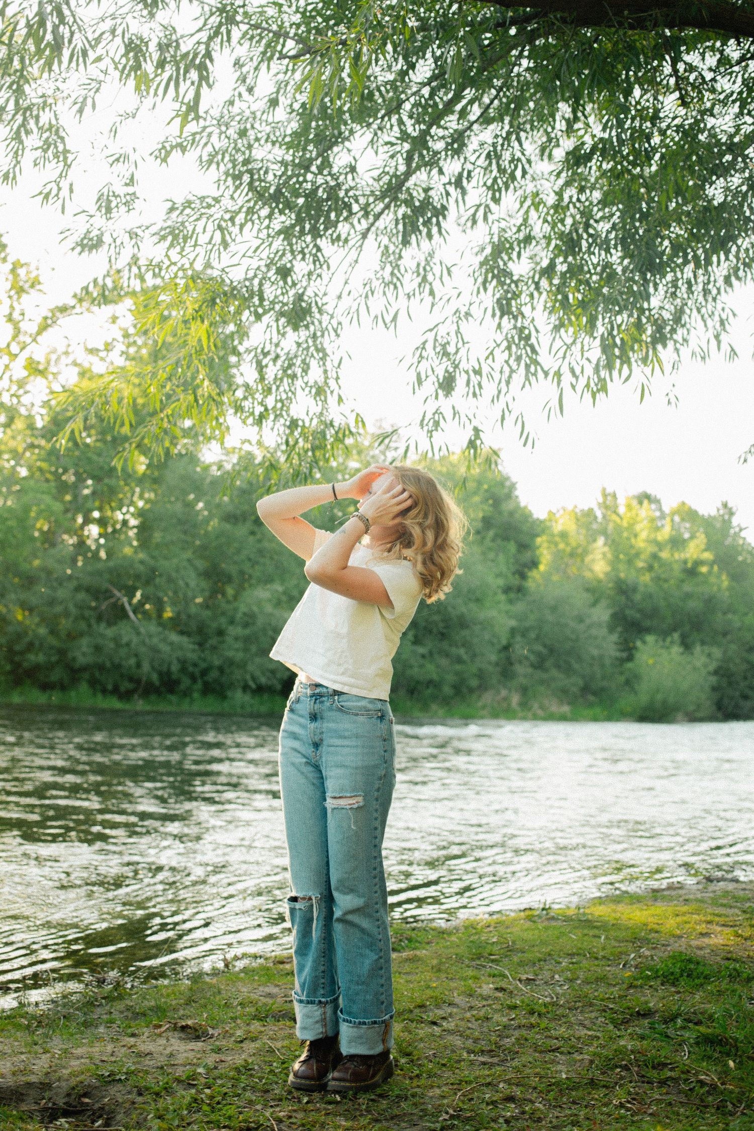 Young woman in white tee and ripped jeans stands by a sunlit river under willow tree branches.