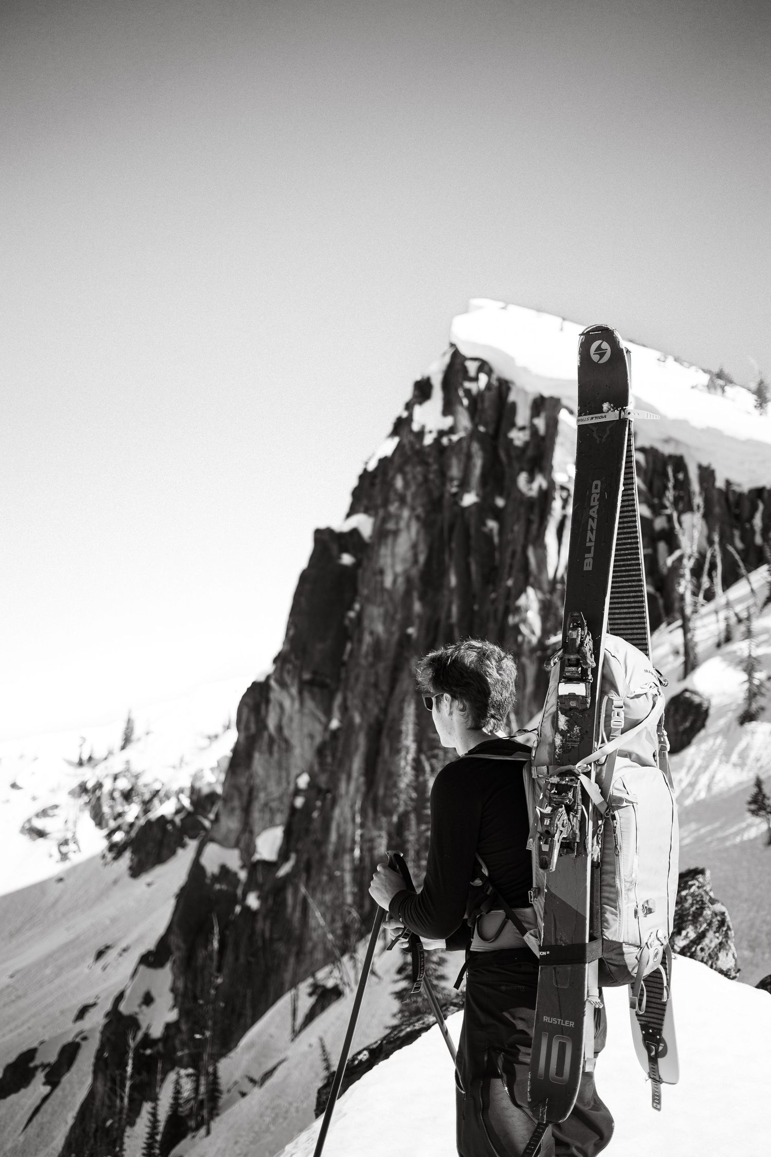Black and white photo of a skier carrying gear on a snowy mountain slope with dramatic rocky cliffs in background in Idaho