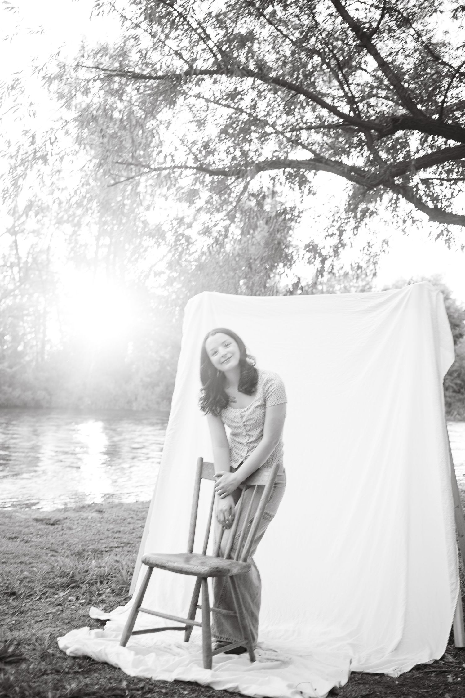 A smiling young woman leans on a wooden chair beside a white backdrop near a sunlit river in black and white.