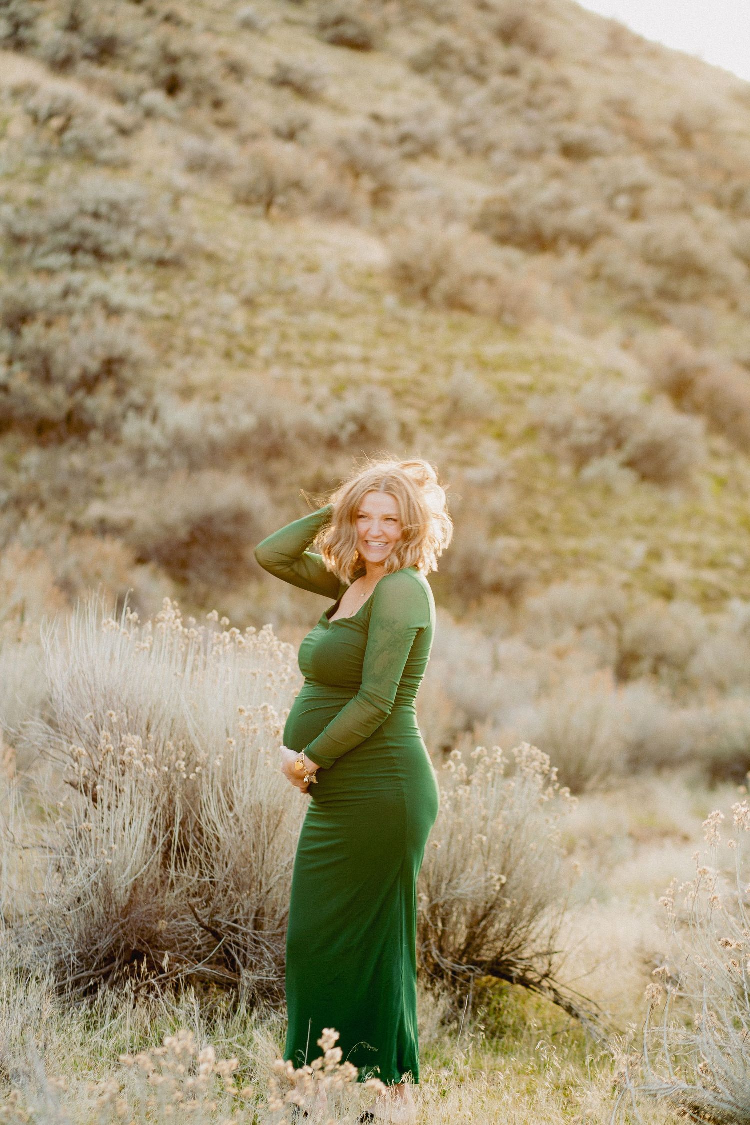 Smiling pregnant woman in green maxi dress poses outdoors among sagebrush with sunlit hillside backdrop.
