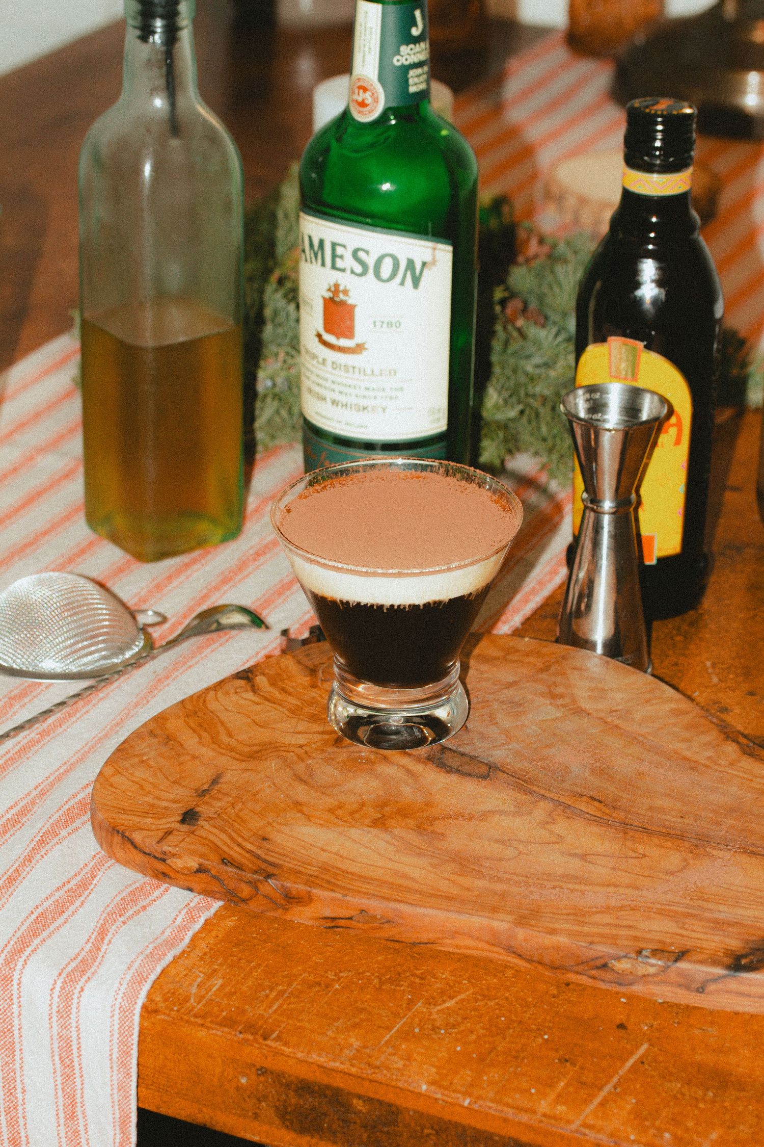 A cocktail with creamy foam sits beside a Jameson whiskey bottle and other liquor bottles on a wooden cutting board taken by stoked media