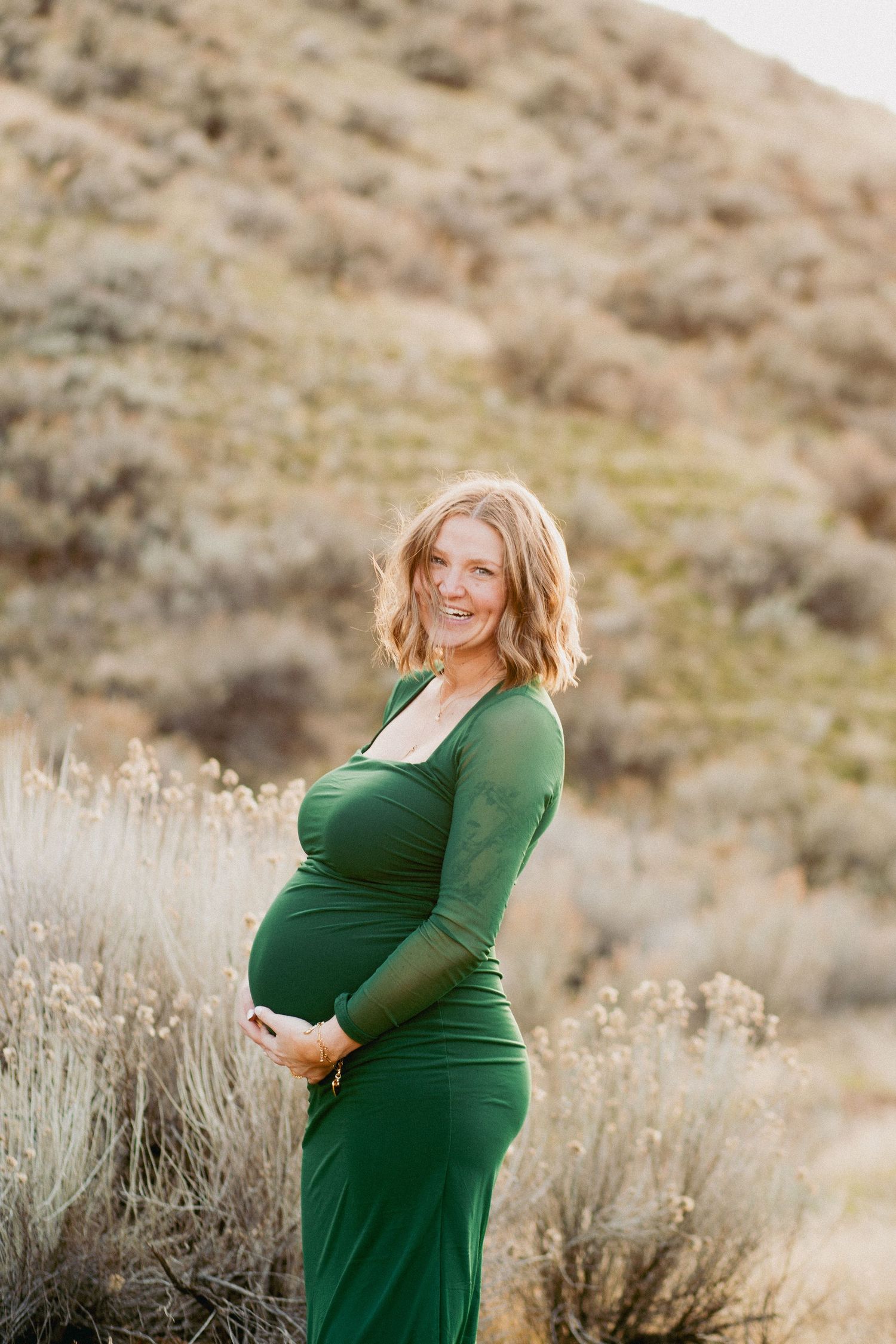 Smiling pregnant woman in green long-sleeve dress posing outdoors with sagebrush hills in background.