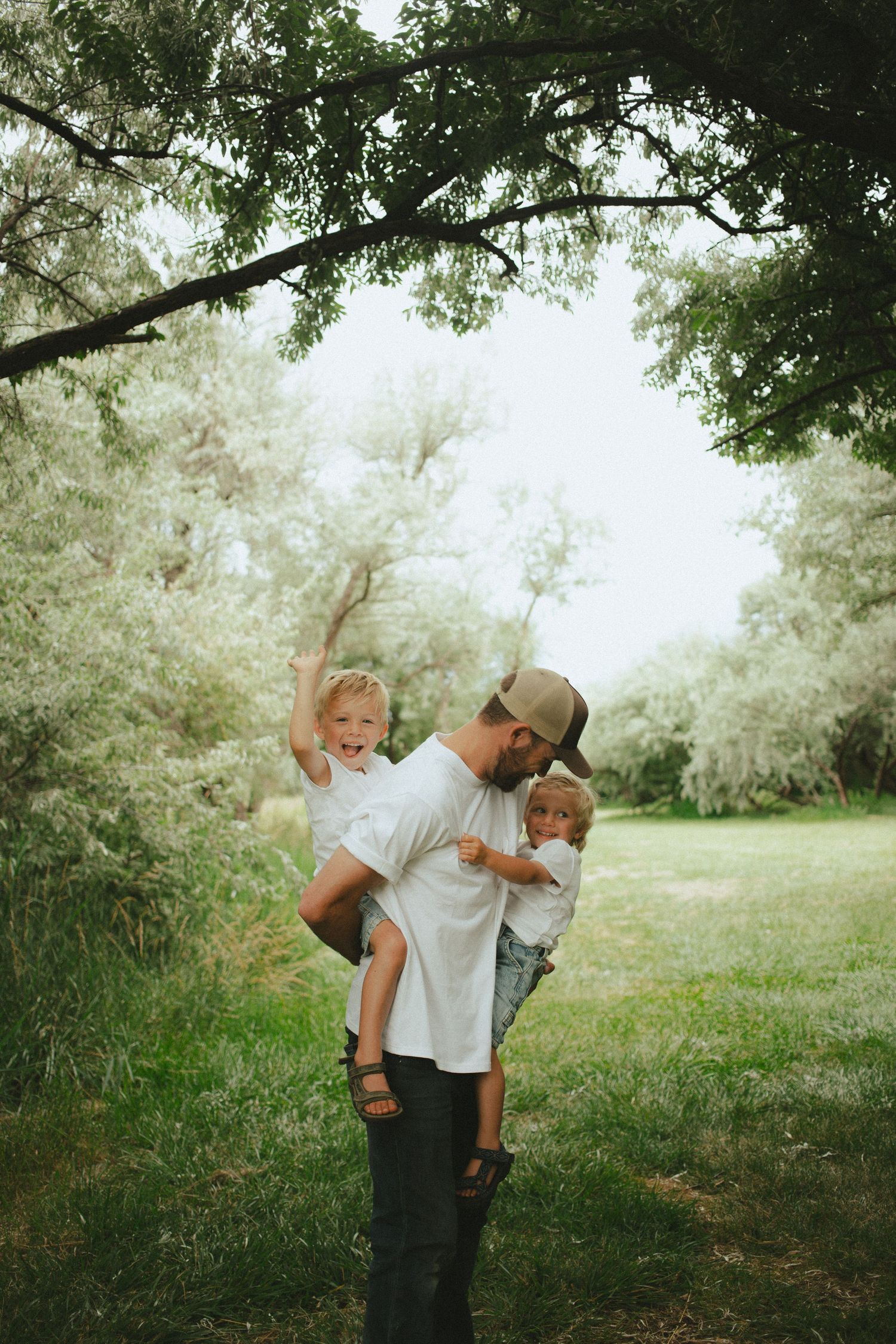 Father holding two young children and laughing under a leafy tree in a lush green outdoor setting.