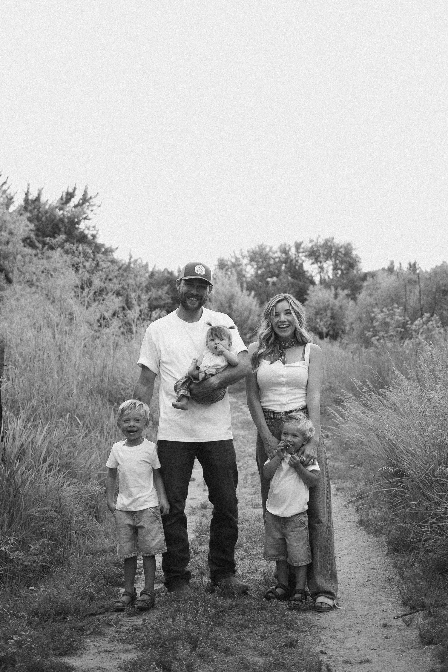 Black and white portrait of family of five posing on nature trail, parents holding newborn baby with two young boys.