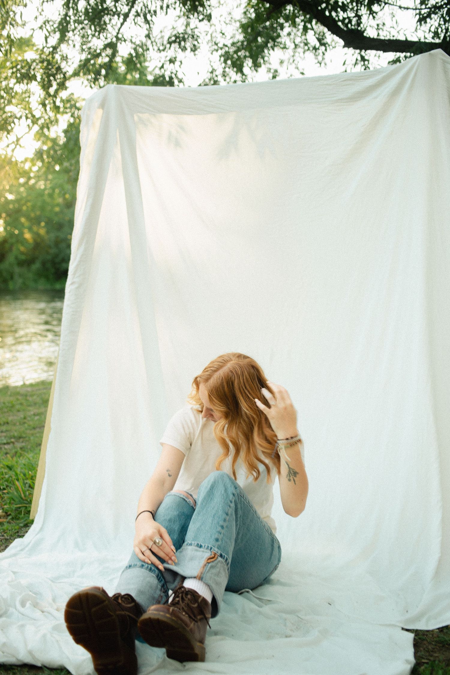Young woman in white tee and ripped jeans sits on white fabric backdrop outdoors near a river.
