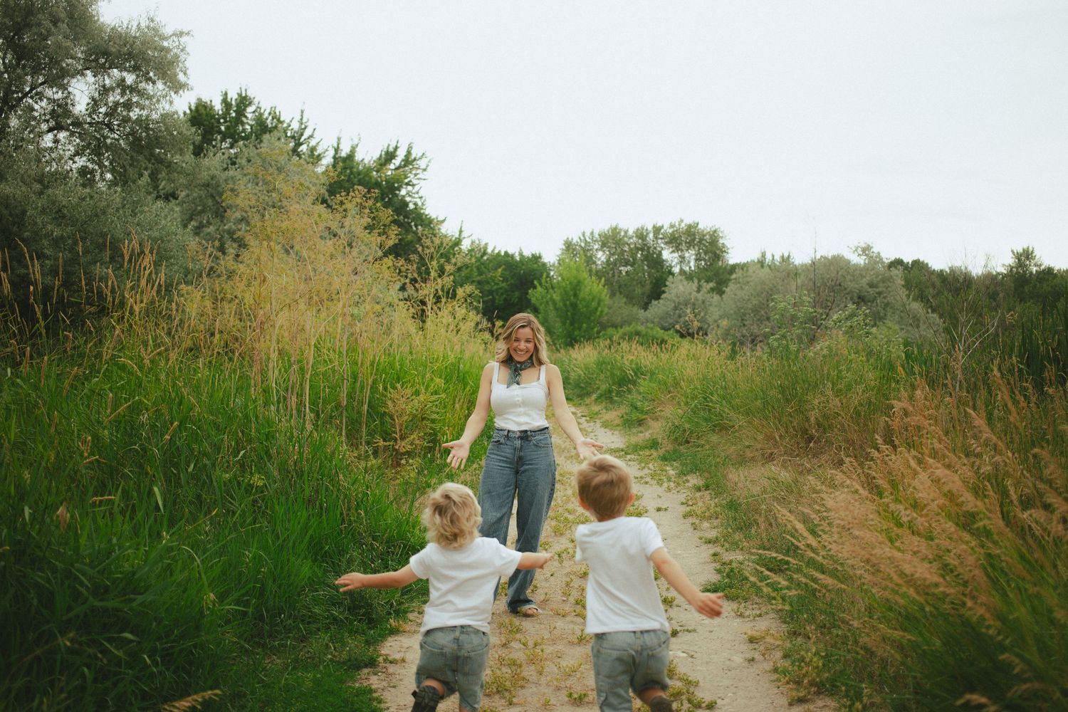 Mother walking with two young blonde toddler boys on a sandy nature trail surrounded by tall green grass.