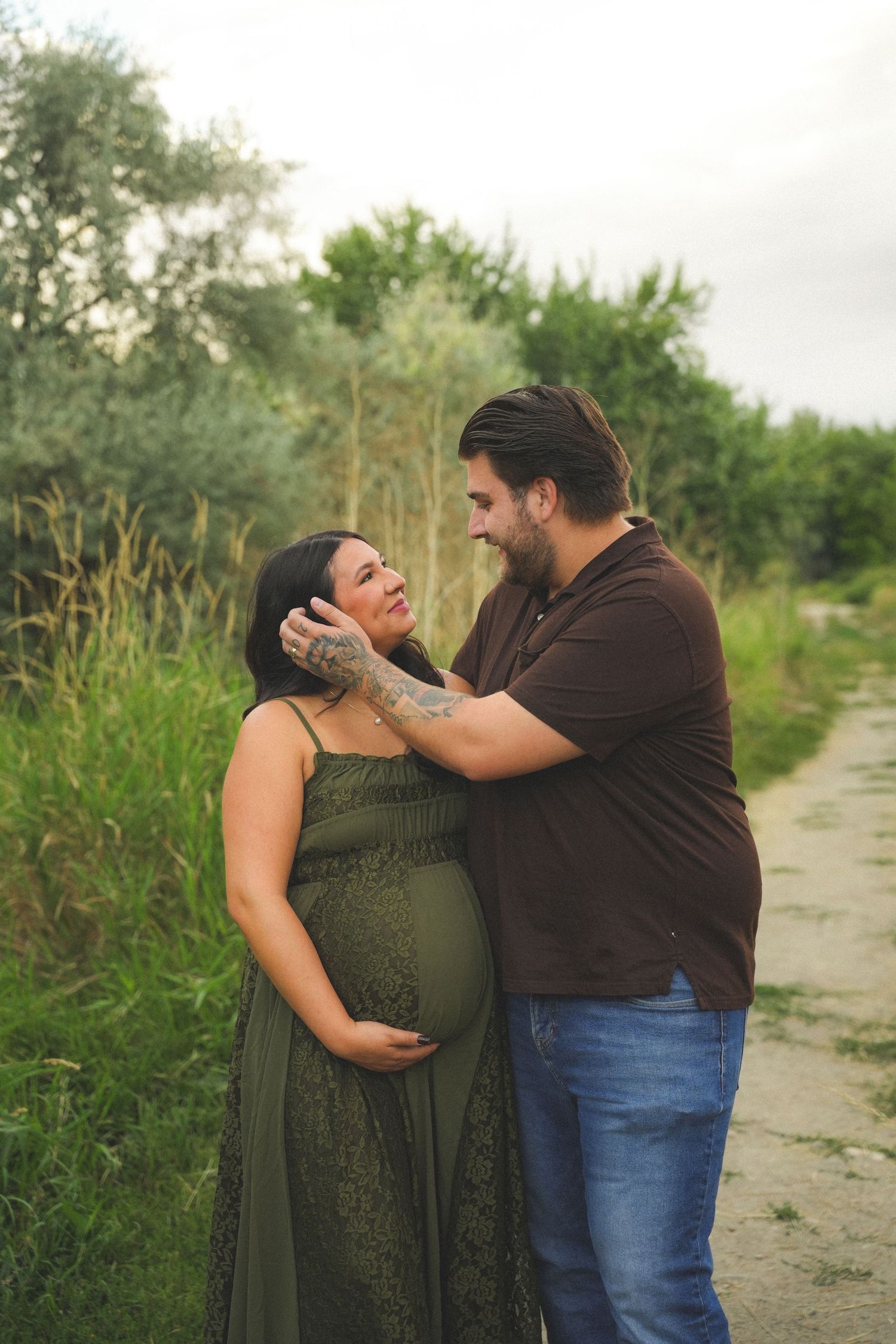 Pregnant couple sharing a tender moment outdoors, woman in green maternity dress, man in brown shirt, lush greenery background.