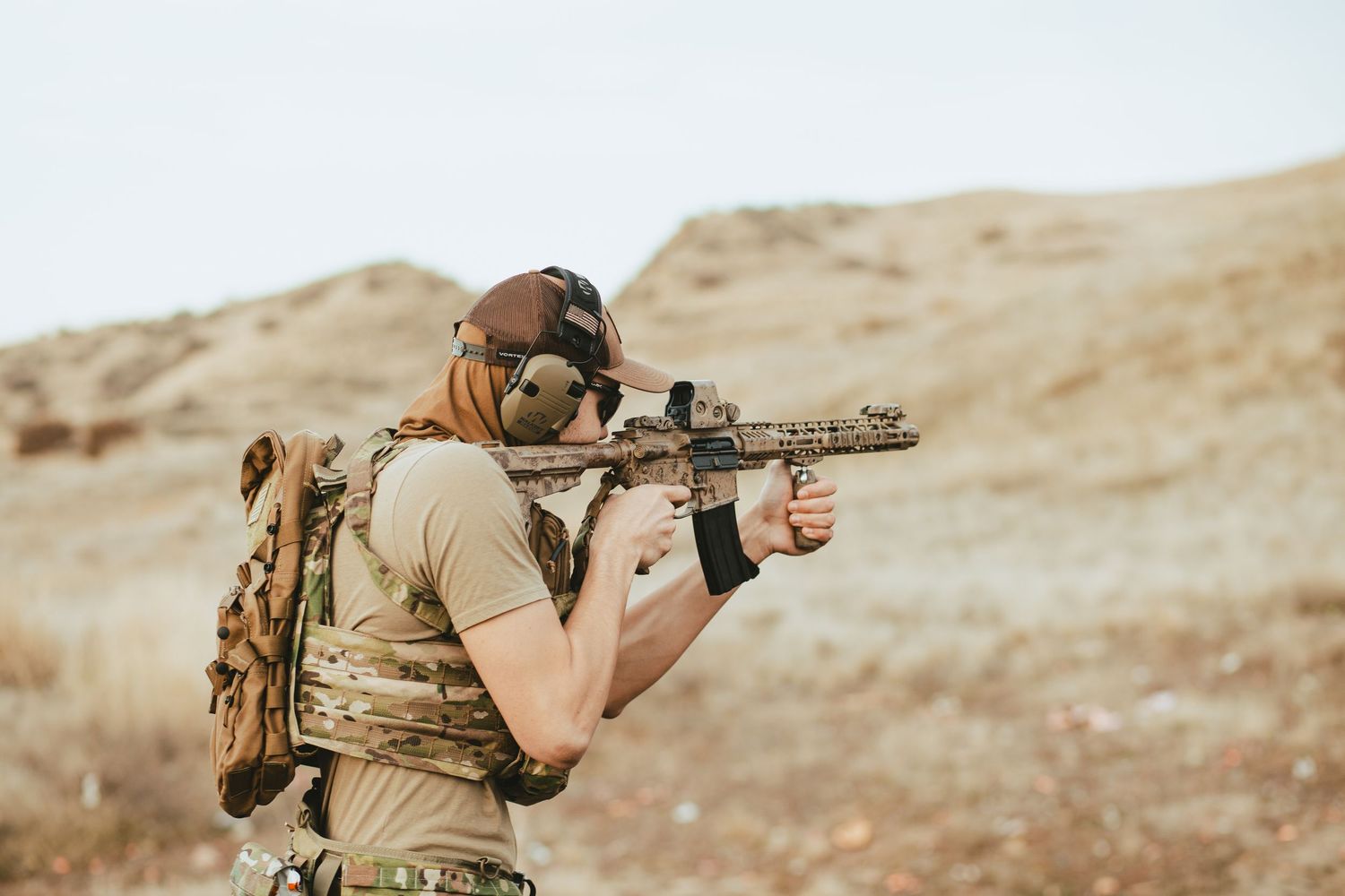 Tactical shooter in desert terrain aims rifle while wearing tan tactical gear and backpack in arid environment in boise Idaho.