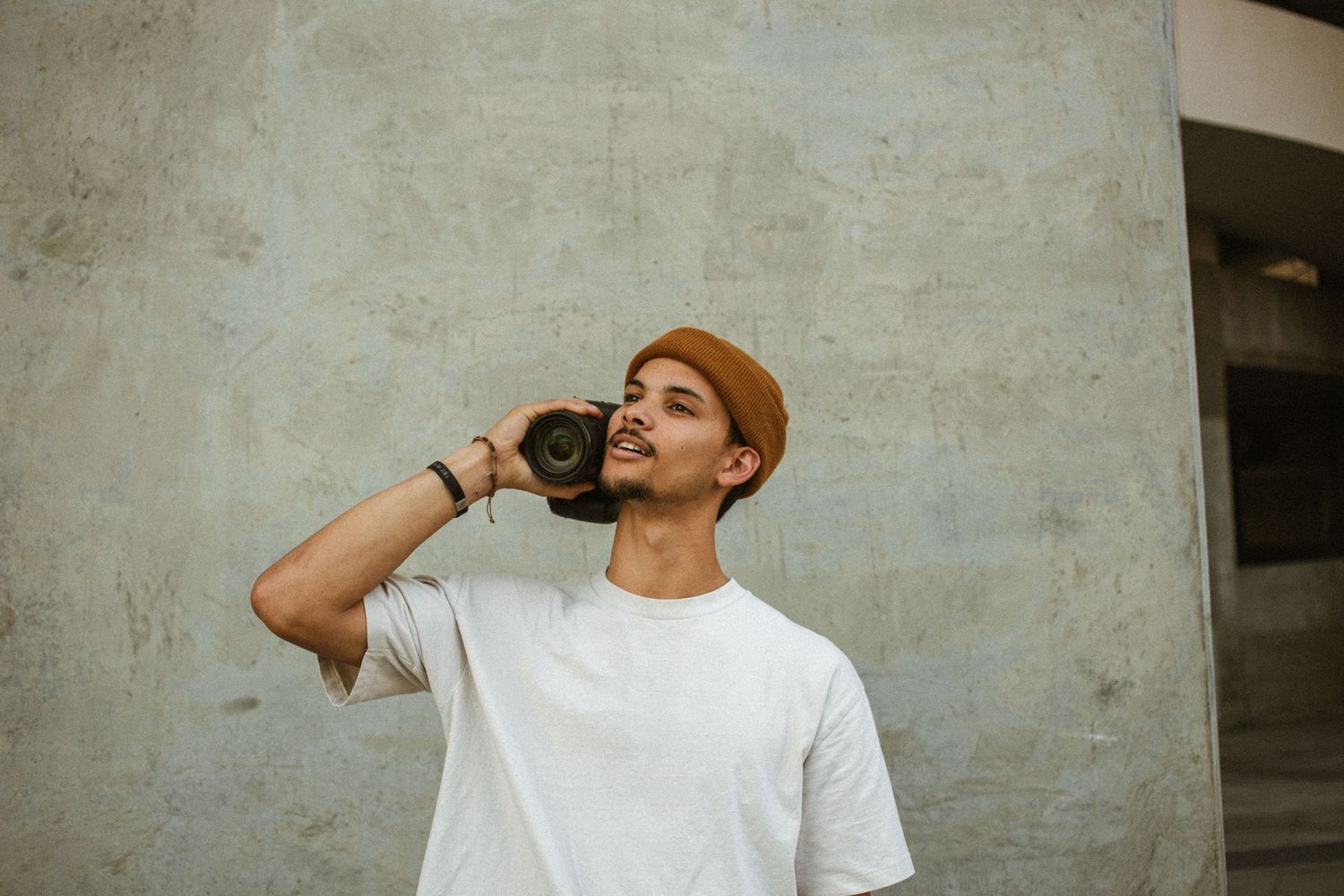 Young man in white t-shirt and brown beanie holding a camera up to his face against a concrete wall.