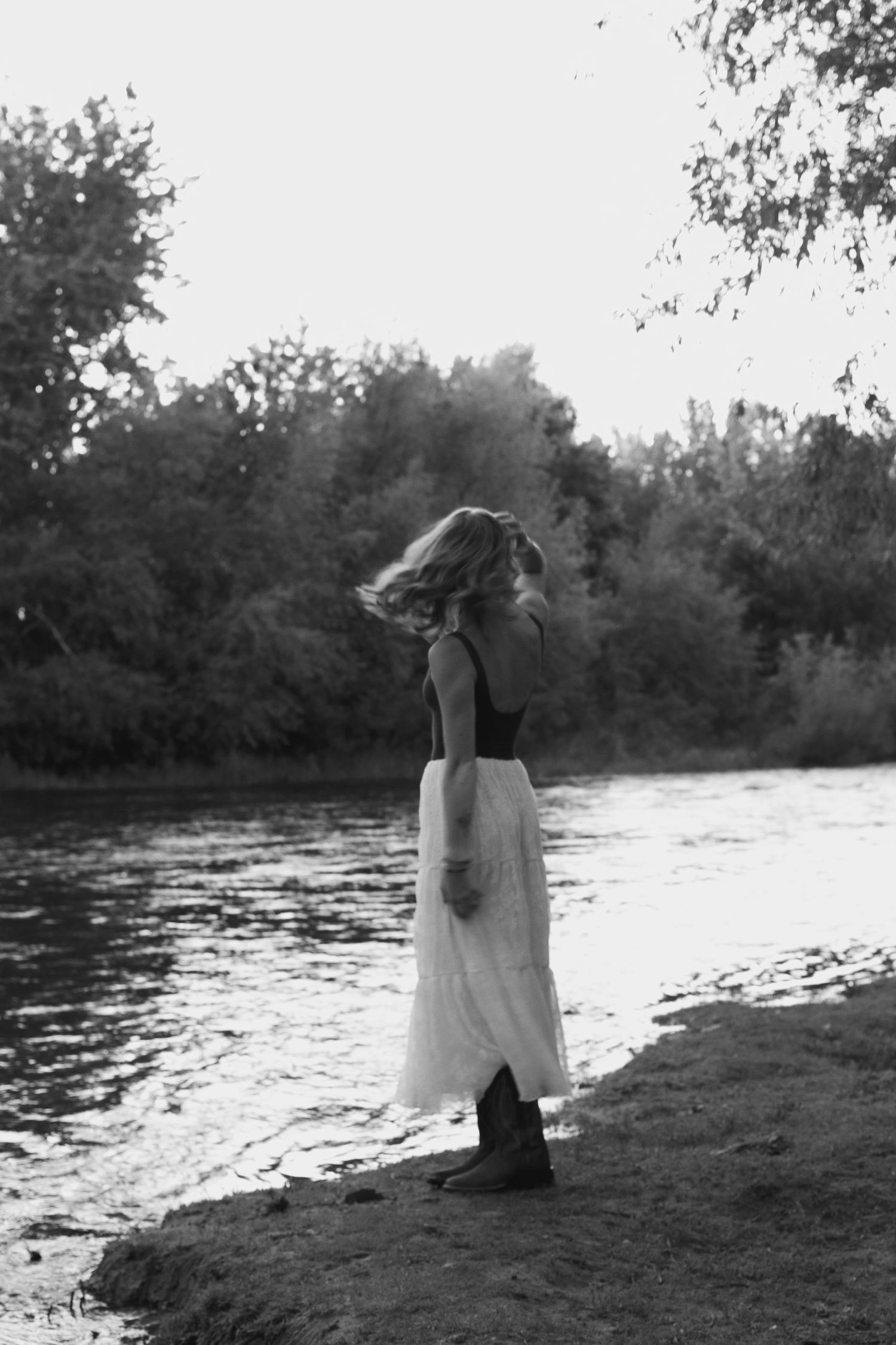 A young woman in a white skirt stands by a river with wind-blown hair in a black and white photo.