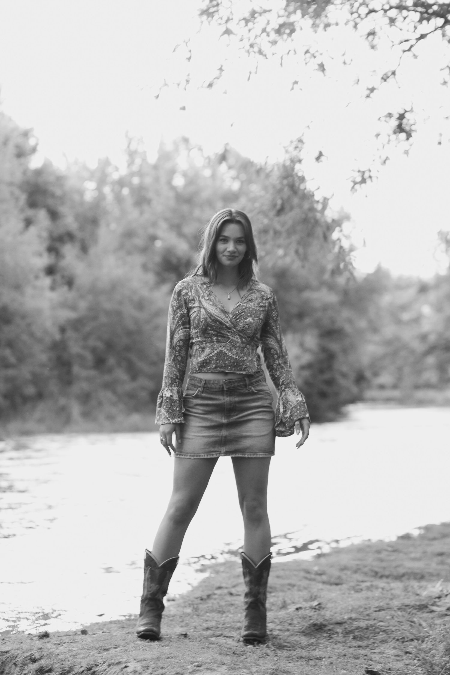 Black and white photo of a woman in a patterned top, denim skirt, and cowboy boots by a riverside.