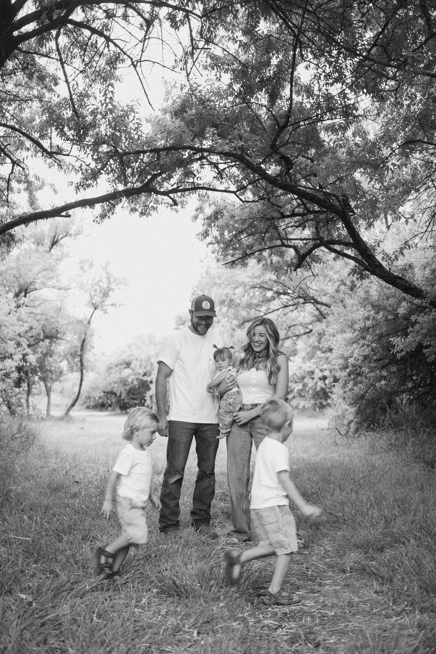 Black and white photo of family of five standing under trees, parents holding newborn while two boys run around.