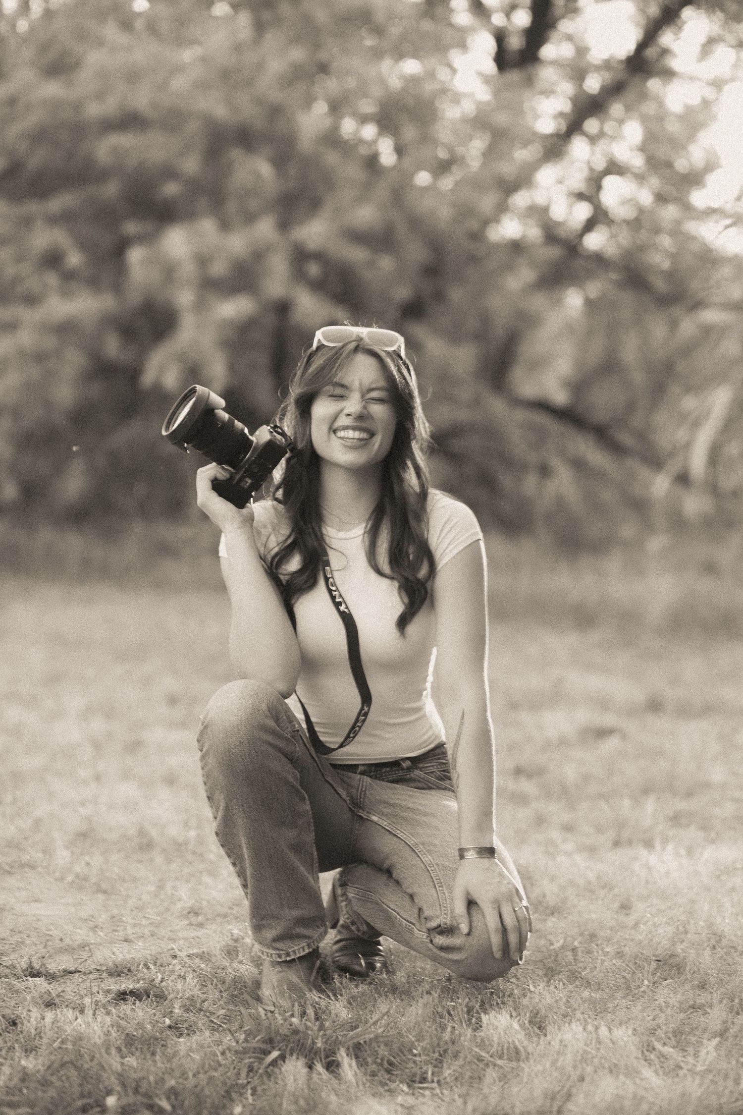 Smiling young woman kneeling on grass holding a DSLR camera in sepia-toned outdoor portrait photo.