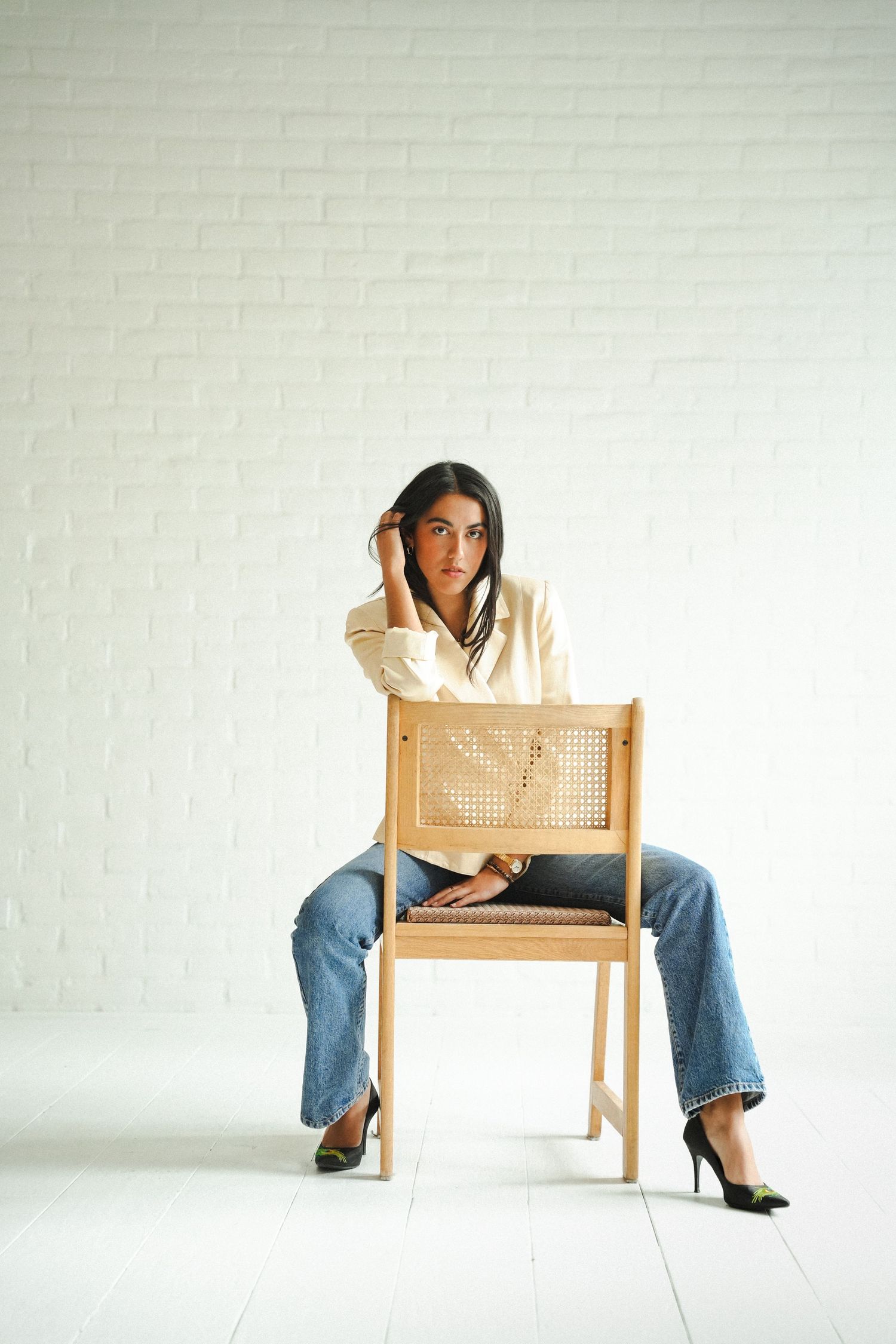 Young woman in white blouse and jeans sitting on wooden rattan chair against white brick wall.