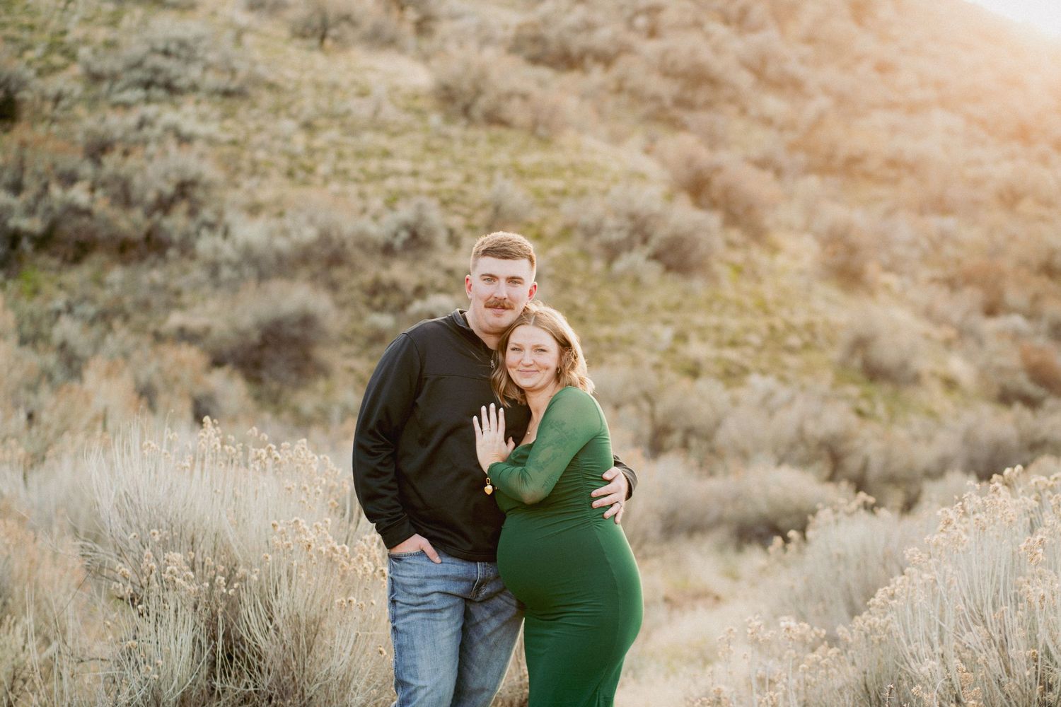 Couple posing outdoors in golden light, woman in green dress showing ring, man in black jacket, hillside backdrop.