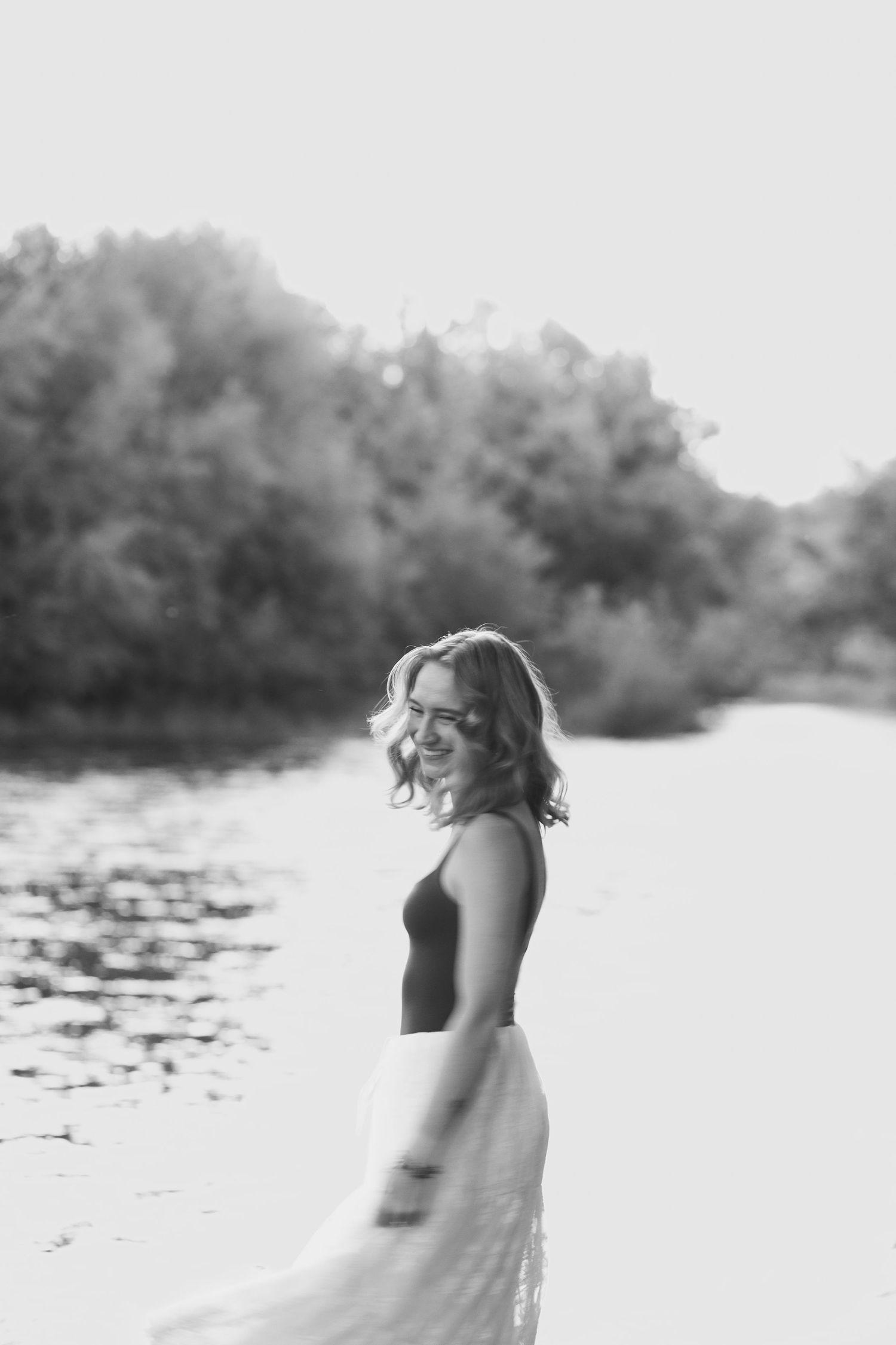 Black and white photo of a laughing woman in a flowing dress spinning on a sunny riverbank.