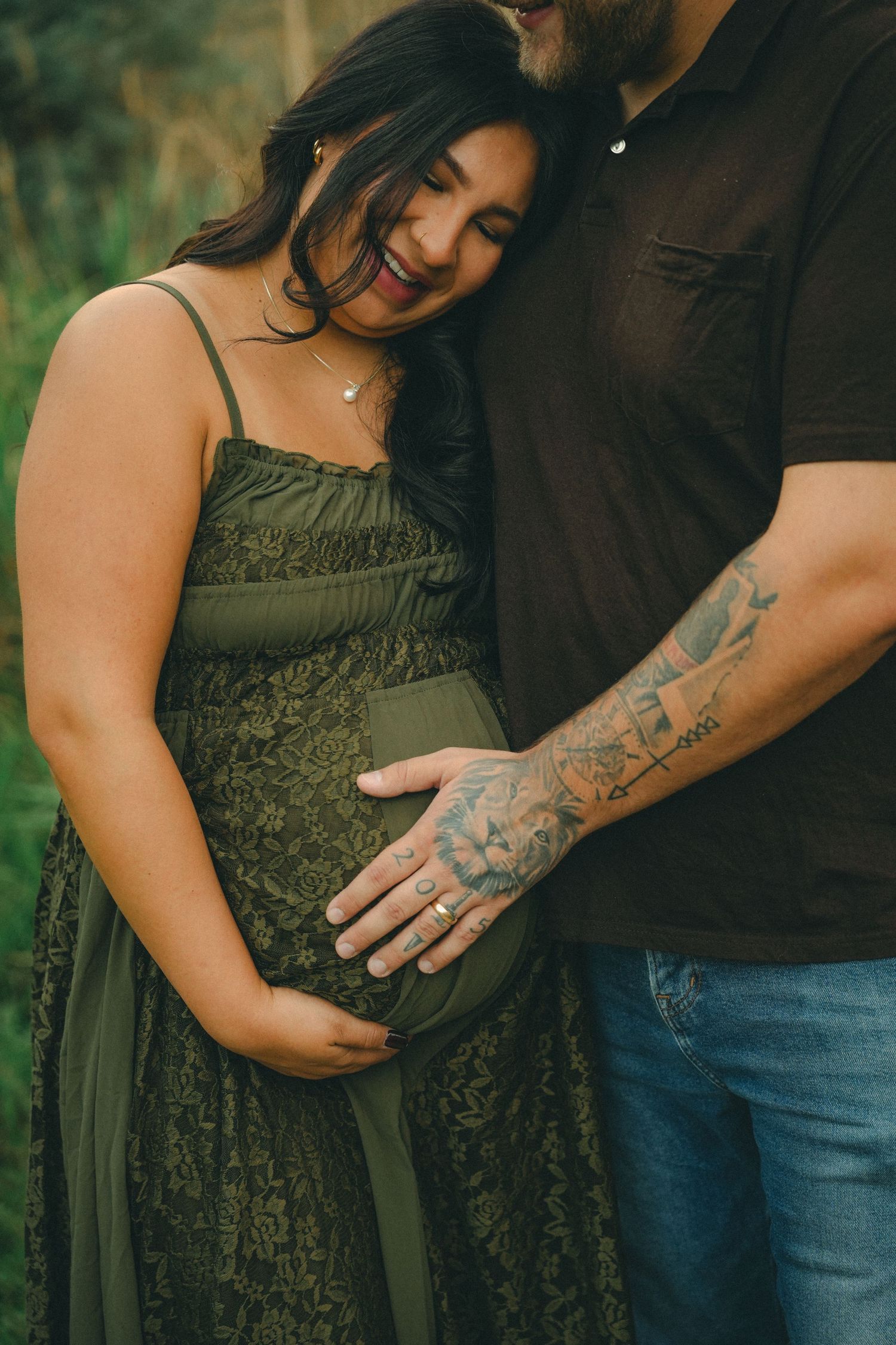 Pregnant couple in green lace dress and tattooed partner tenderly cradling baby bump outdoors.