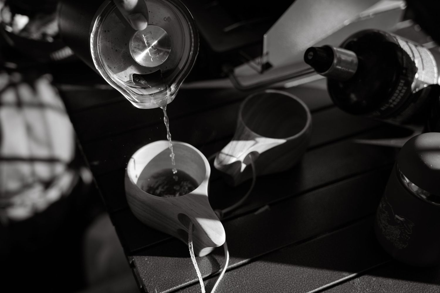 Black and white photo of water being poured into a white cup on a dark table with a wine bottle nearby.
