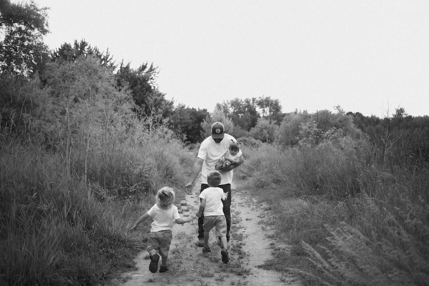 Black and white photo of two young boys running toward their father holding a baby on a rural dirt path.