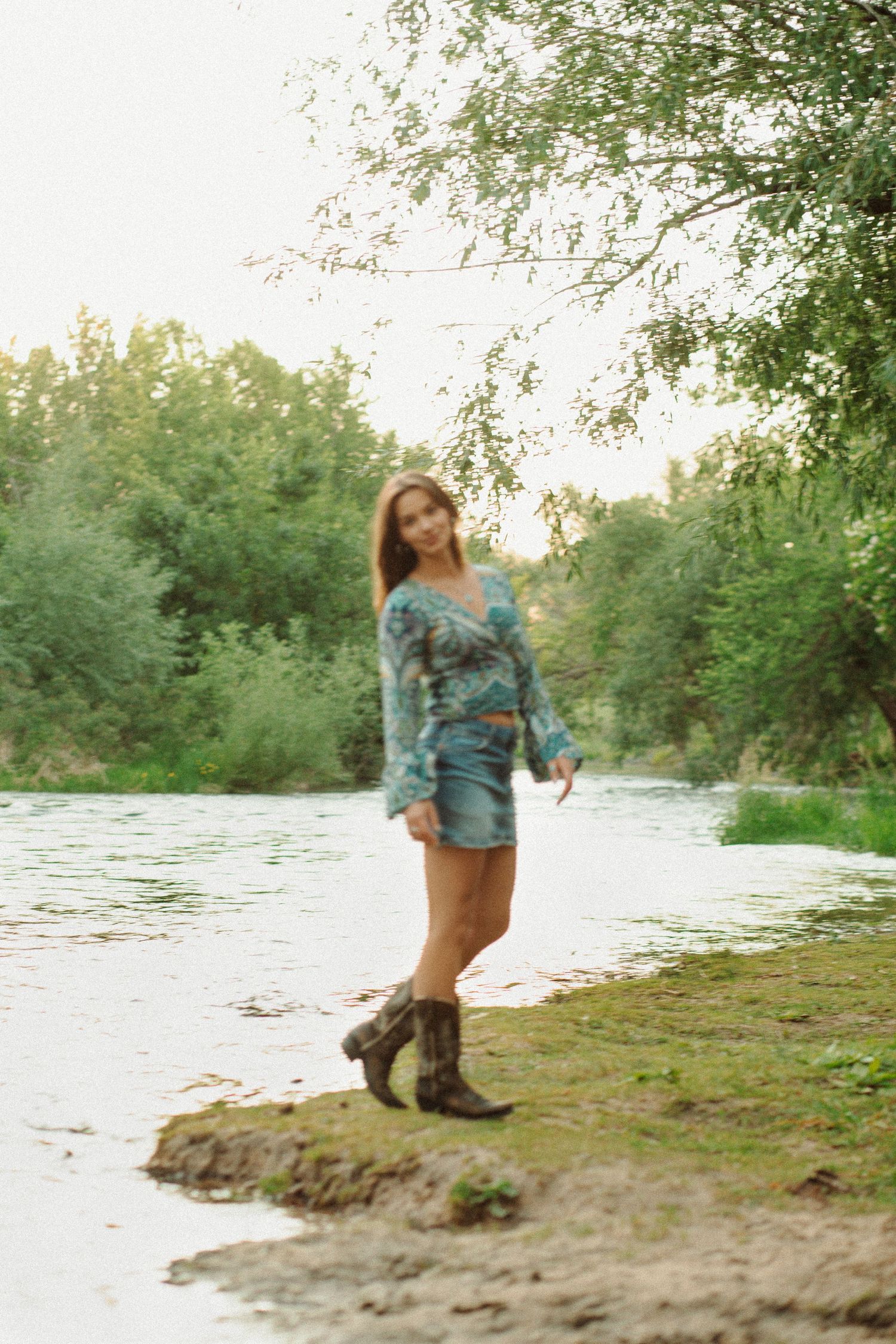Woman in teal floral wrap top, denim mini skirt, and cowboy boots posing by a tree-lined river.