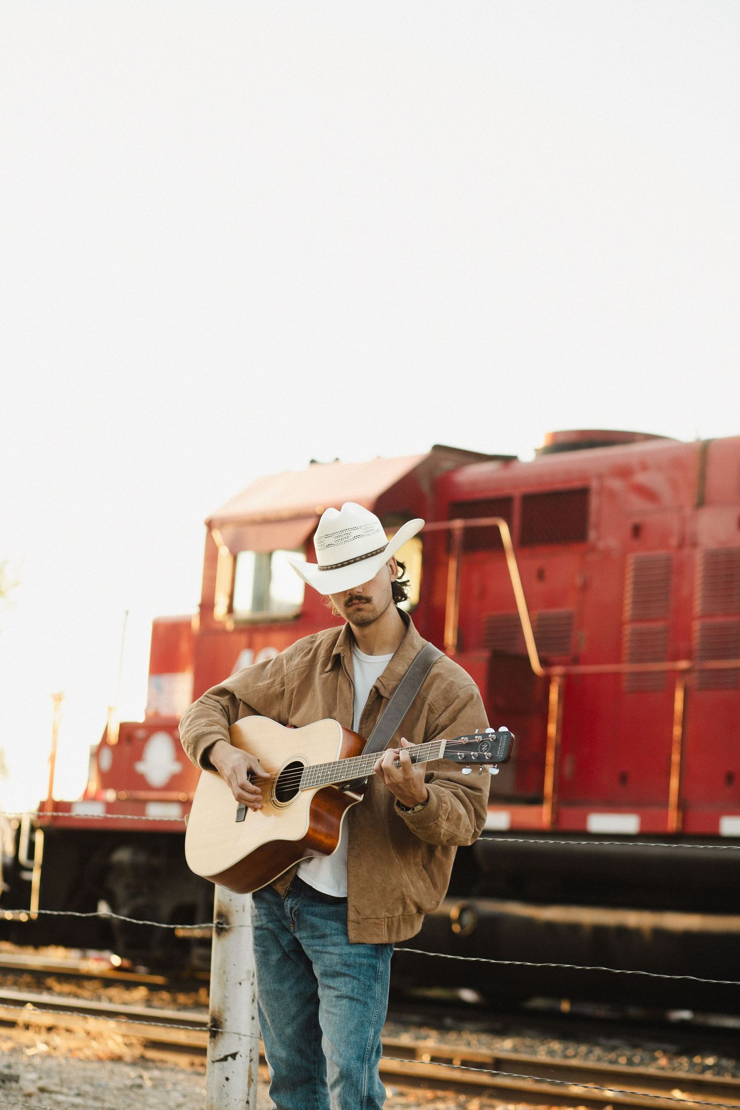 Colorful image show A young man in a cowboy hat and jeans playing his guitar in front of a red train passing by in Emmett Idaho