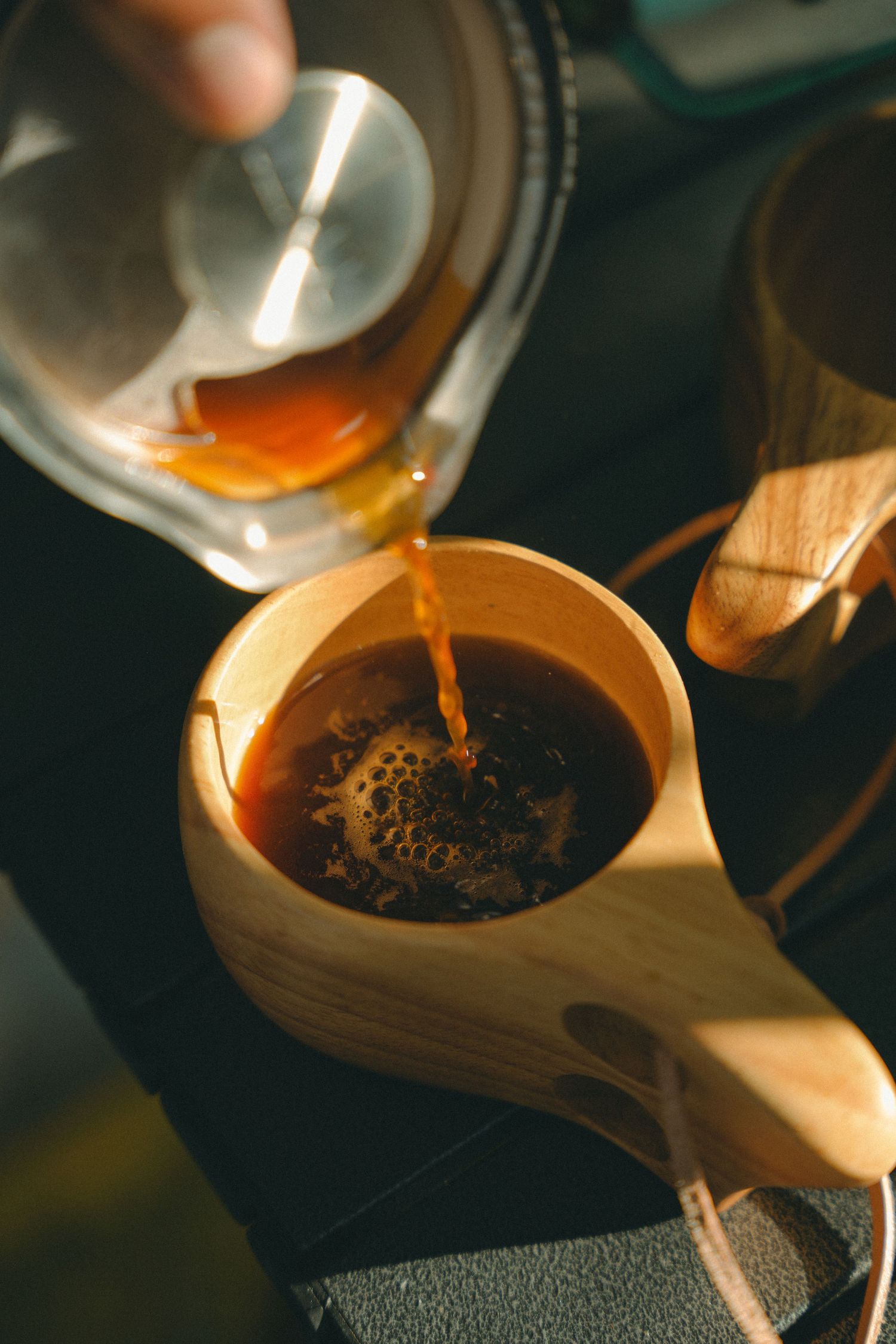Honey being poured from a glass pitcher into a wooden cup of coffee in warm natural light.