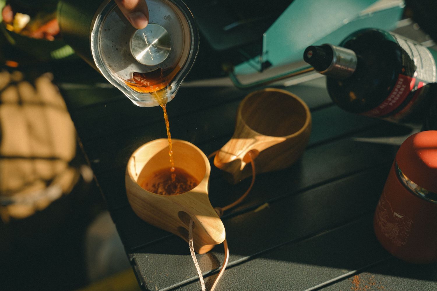 Hot tea being poured from a glass kettle into a wooden cup on a dark outdoor table.