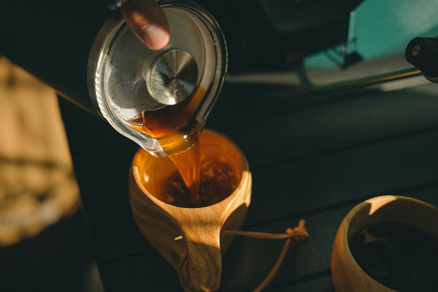 Hot tea being poured from a glass teapot into a wooden cup on a dark surface.