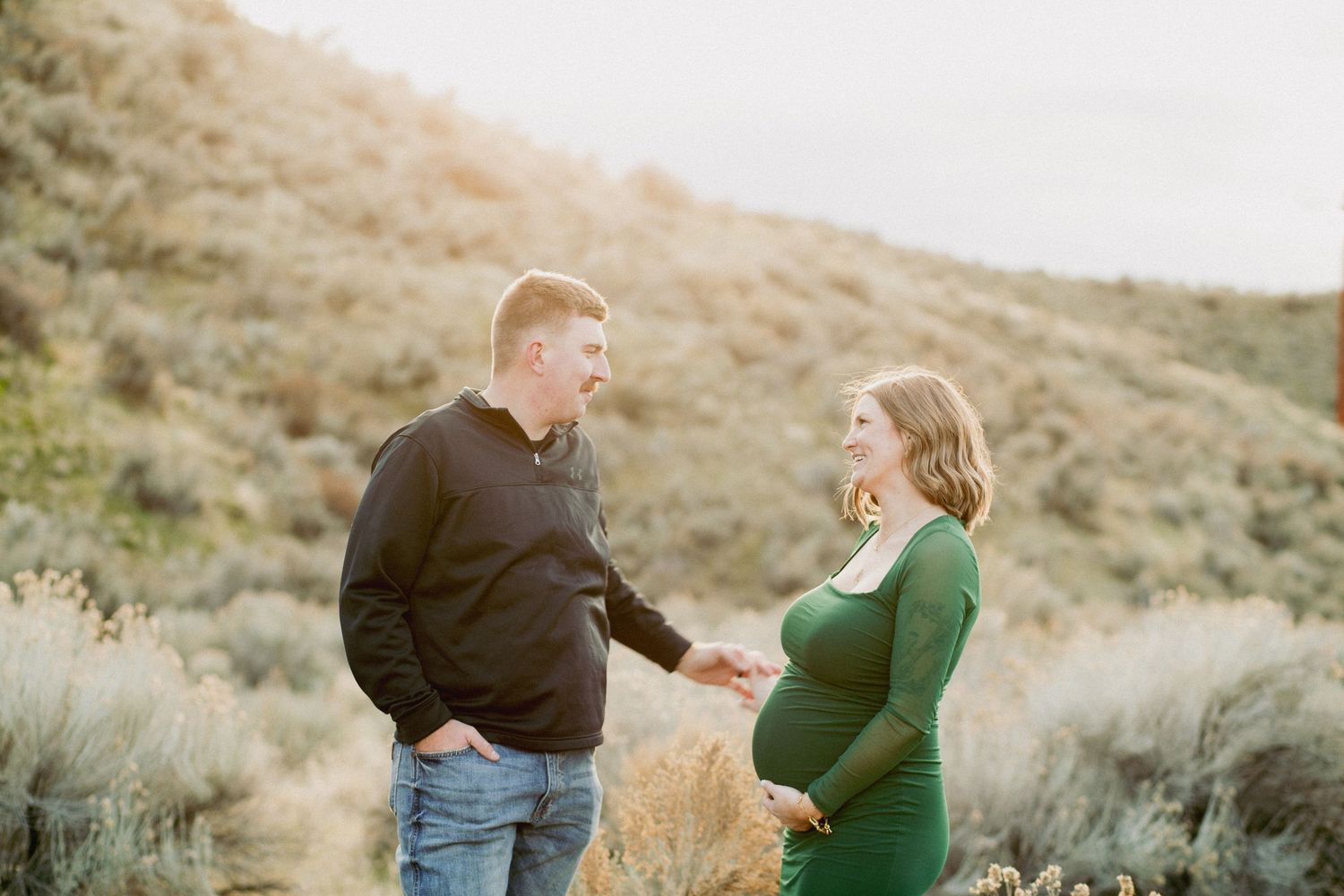 Pregnant couple holding hands outdoors in a sunny desert landscape, woman in green dress, man in black hoodie and jeans.
