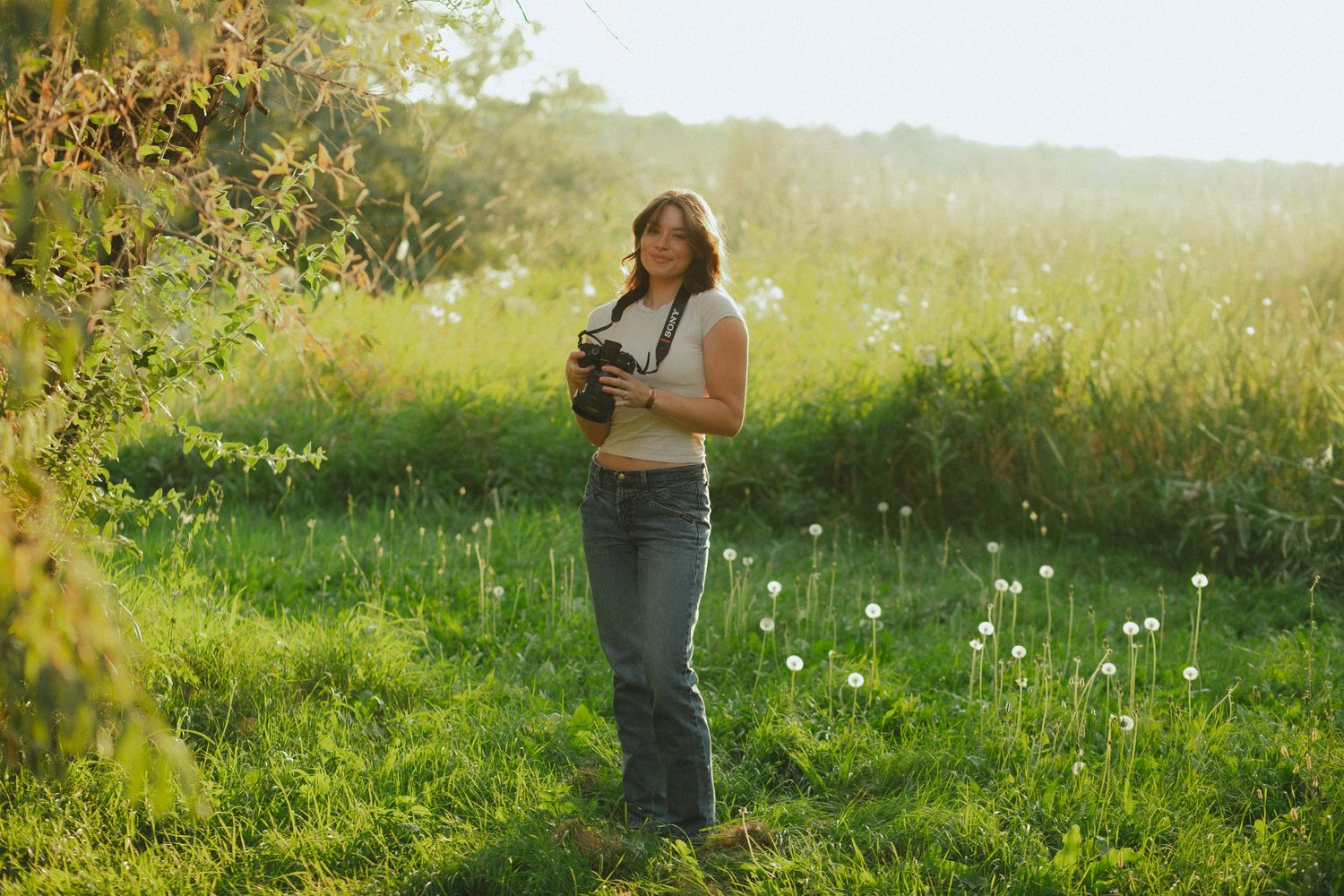 Woman photographer standing in sunny green meadow holding camera, surrounded by wildflowers and lush grass in golden light.