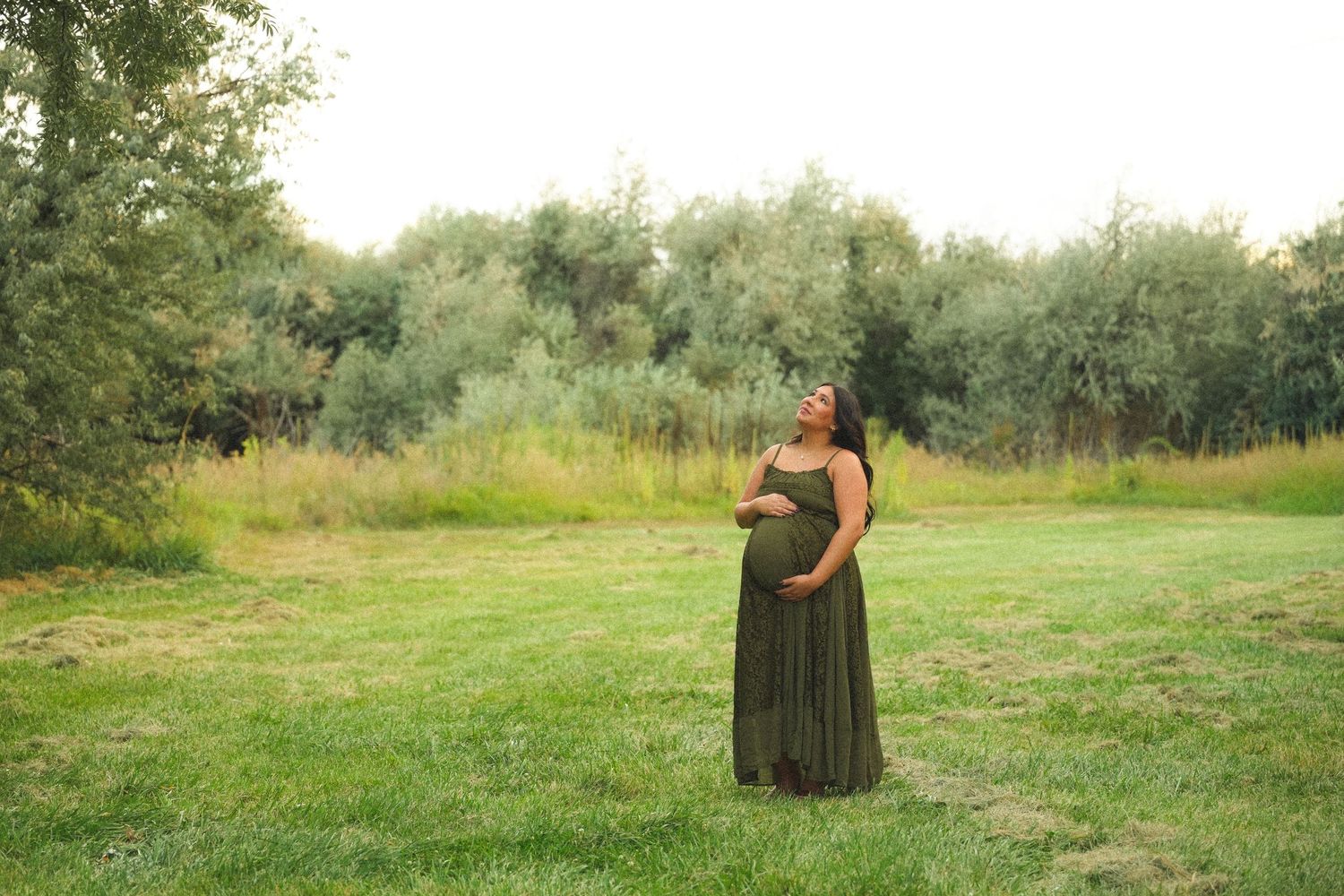 Pregnant woman in olive green maxi dress standing in lush green field, cradling baby bump while gazing upward.