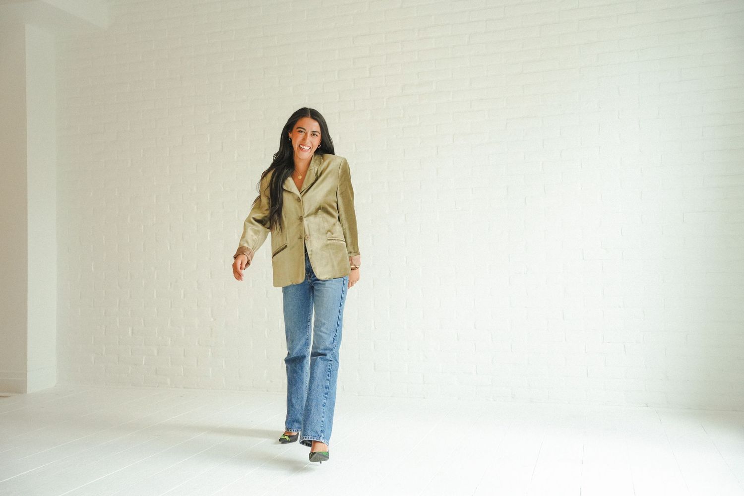 Woman in olive blazer and blue jeans smiling in bright white room.