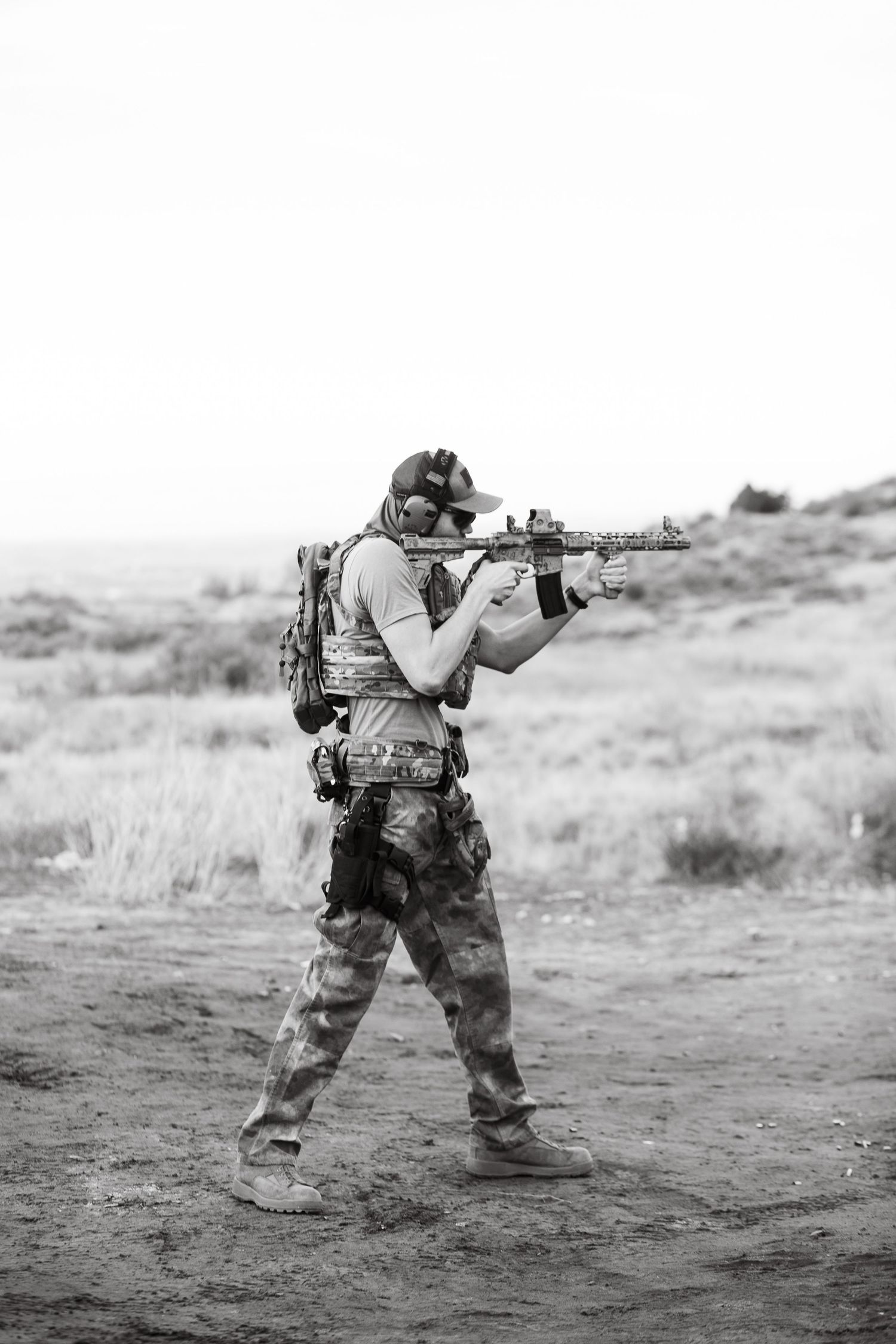 A soldier in desert camouflage crouches in a combat stance while aiming a rifle across an arid landscape in the boise Idaho hills