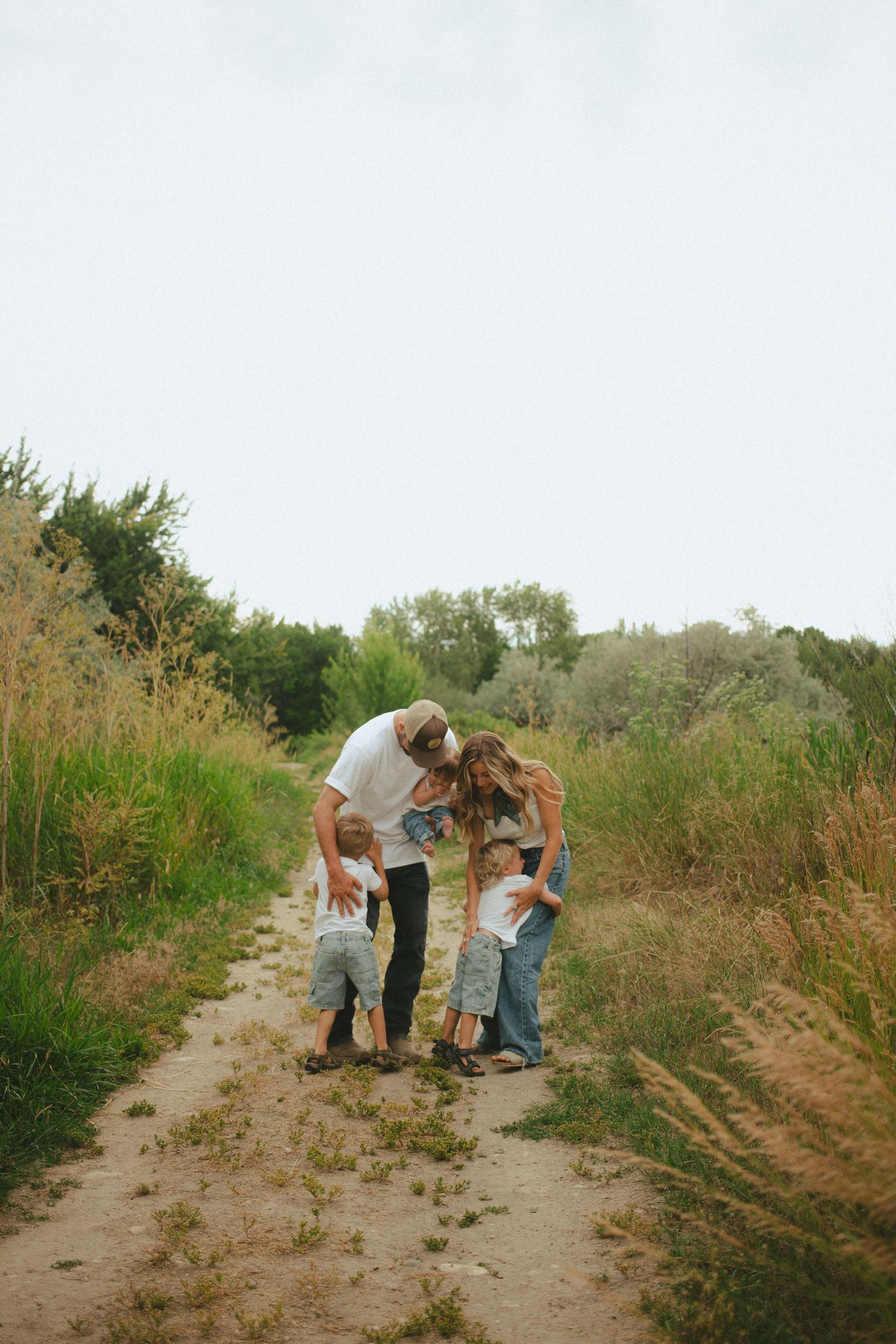 A loving family of five huddles together on a rustic dirt trail surrounded by lush green foliage.