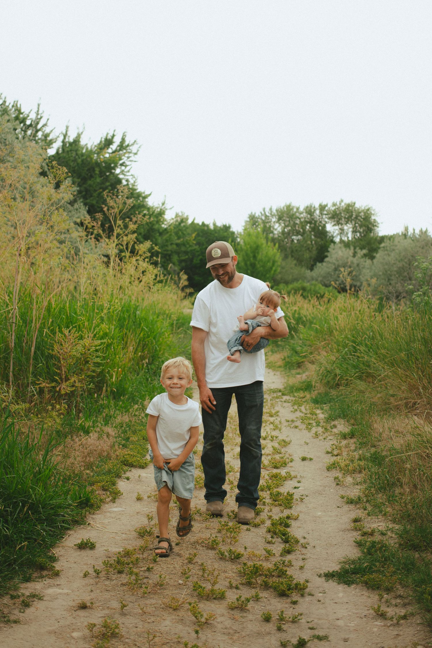 Father in white tee and cap walks sandy trail holding newborn baby while young blonde son runs beside him.