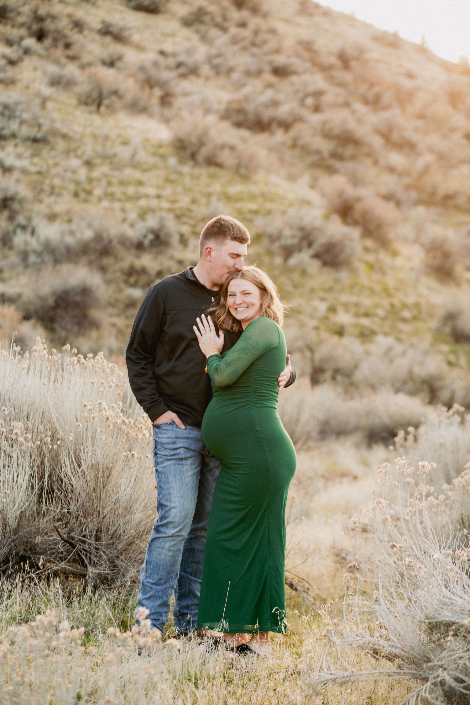 Couple embracing outdoors in sagebrush landscape, woman in green dress, man in black sweater and jeans, golden hour light.