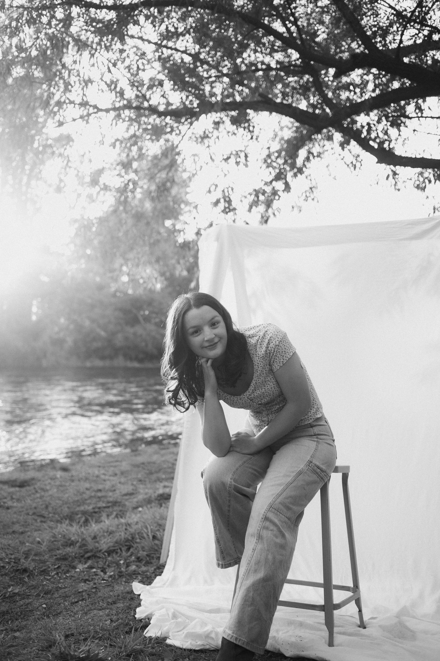 Black and white portrait of a woman sitting on a stool outdoors near a white fabric backdrop by a river.