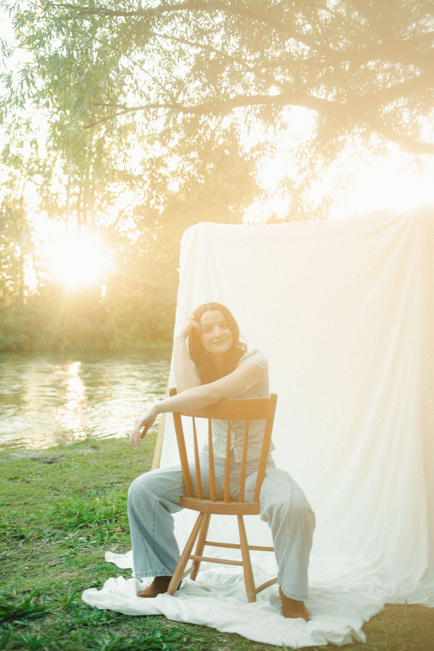 A cheerful woman sits on a wooden chair beside a white backdrop bathed in warm golden sunset light outdoors.