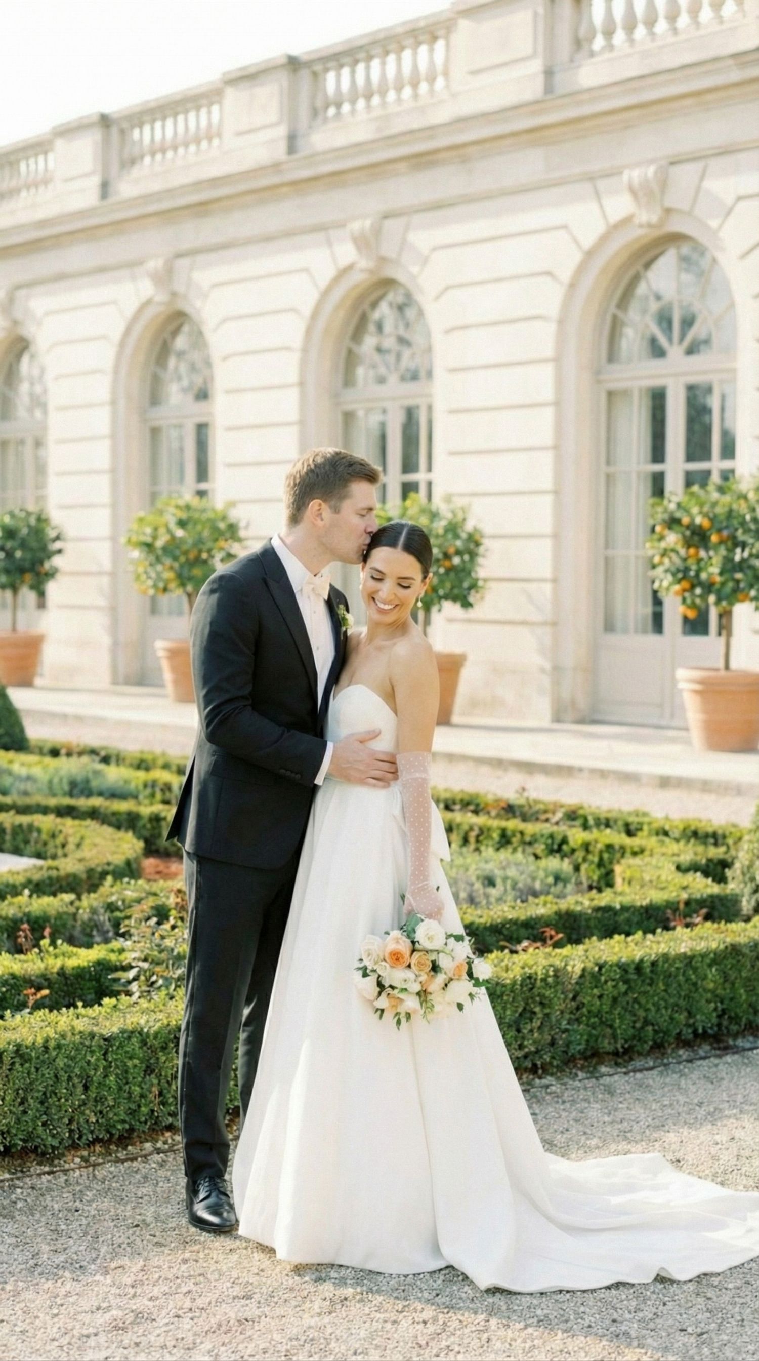 Newlyweds share intimate moments in a formal garden setting with elegant architecture and manicured hedges in the background.