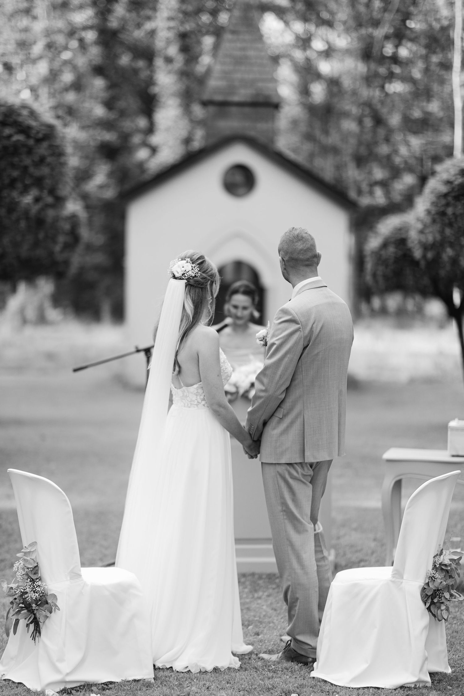 Black and white outdoor wedding ceremony setup with white chairs and a small chapel in the background. Luxembourg wedding photographer