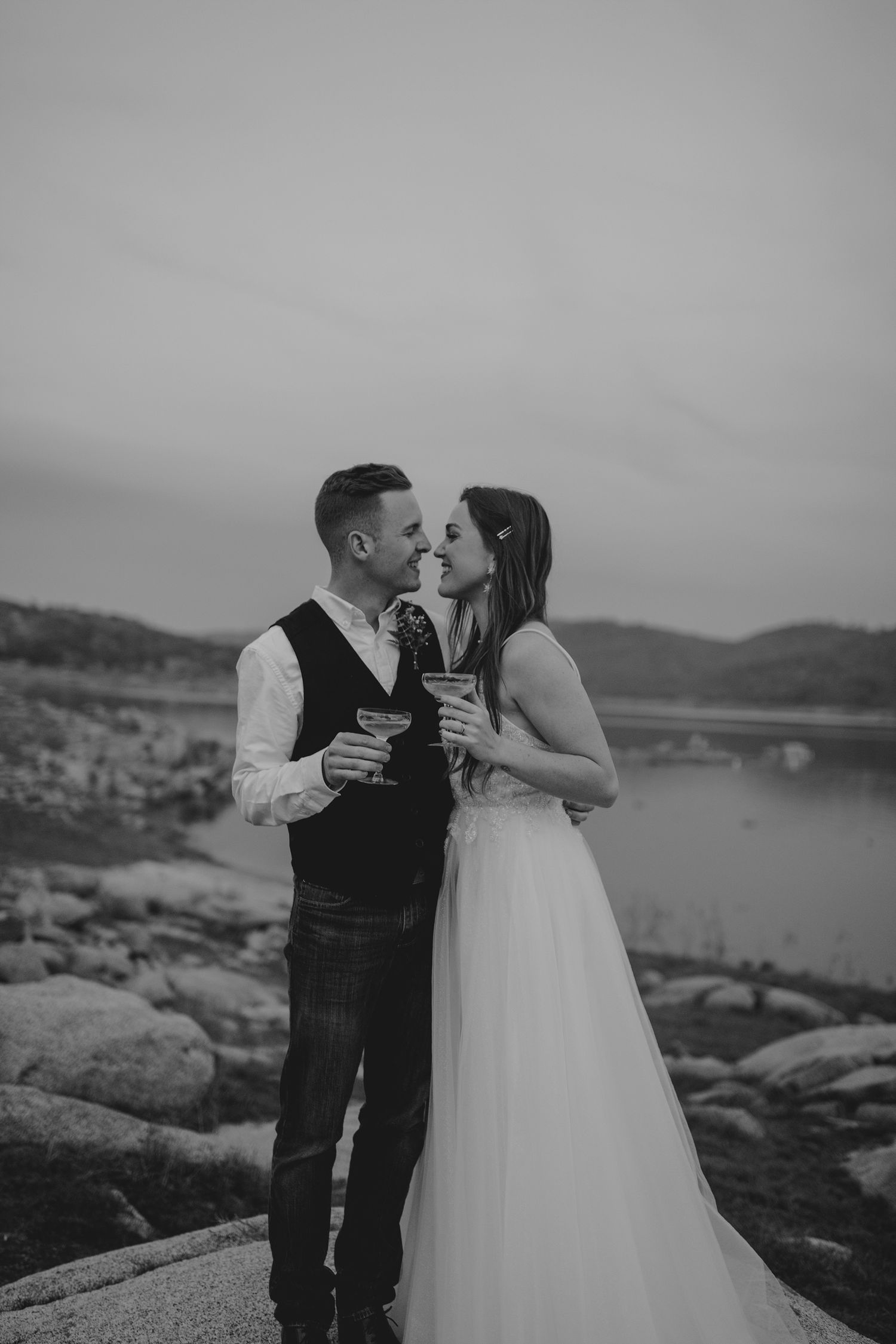 A romantic black and white wedding photo of a couple toasting with drinks on rocky coastal cliffs at sunset.
