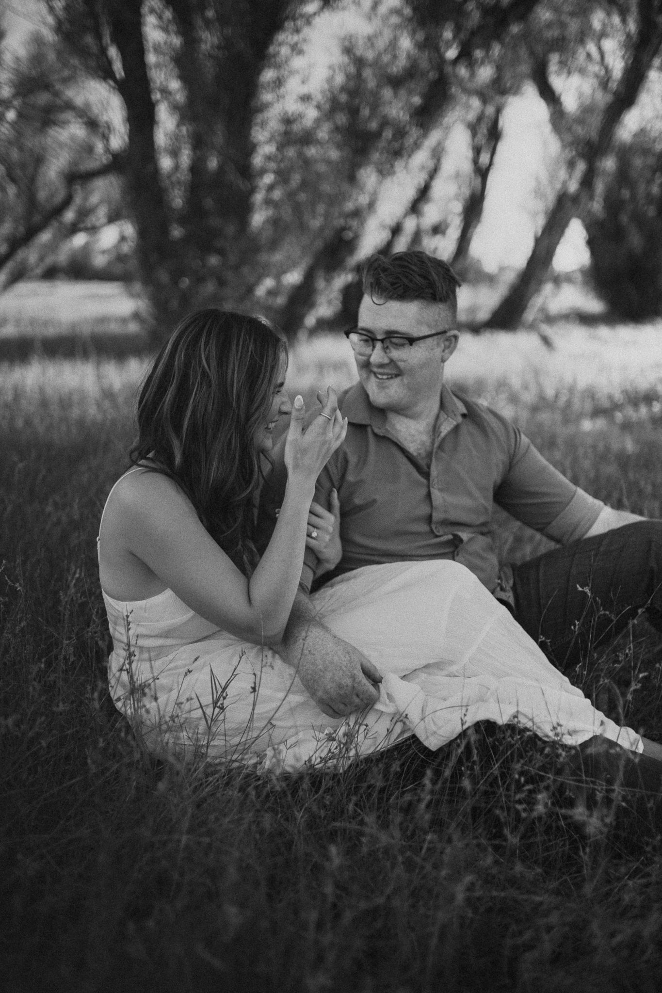 A romantic black and white portrait of a couple in Folsom sitting together in a grassy meadow with trees in the background.